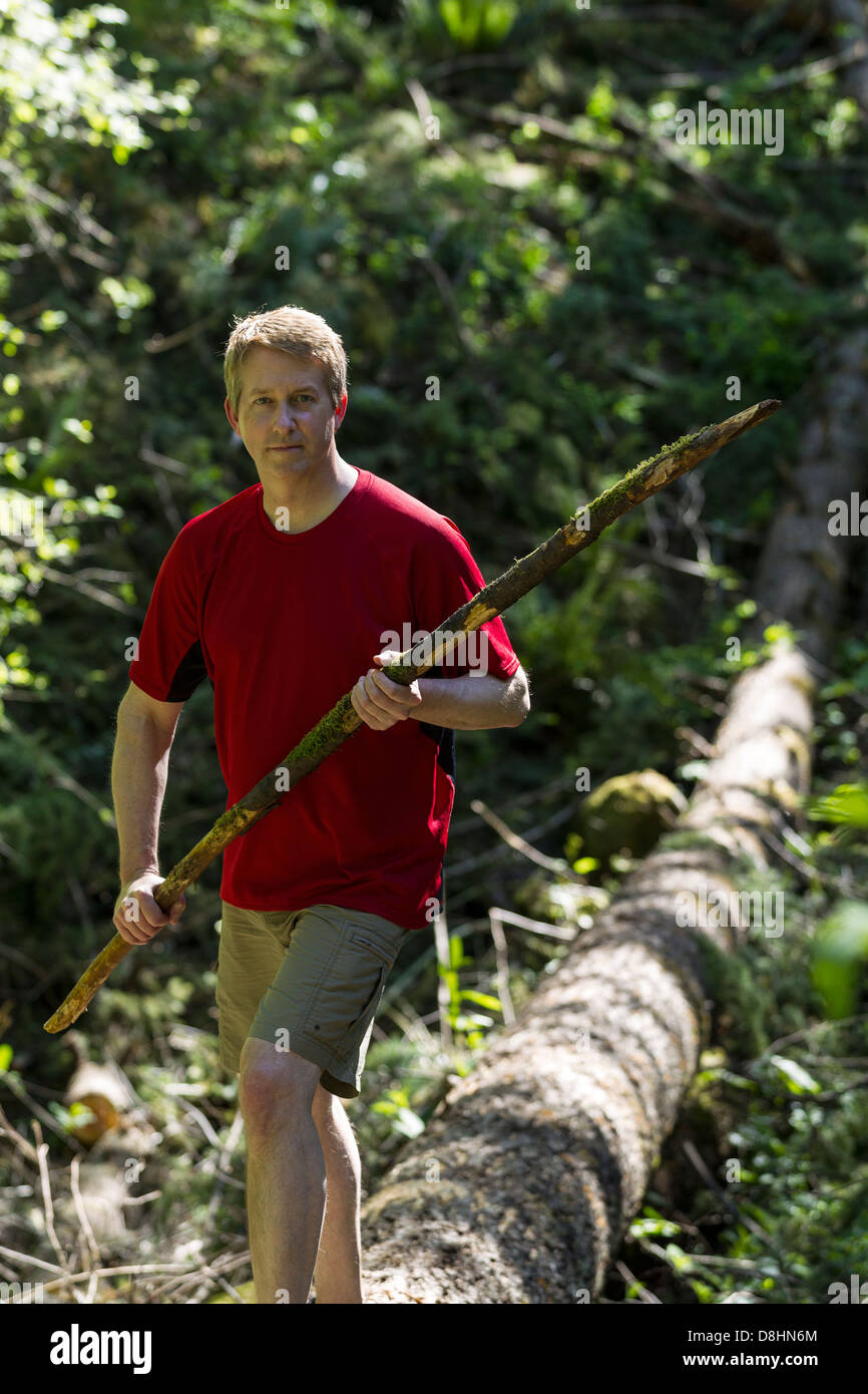 Vertical photo of mature man crossing over a fallen tree while holding a walking with trees in background Stock Photo