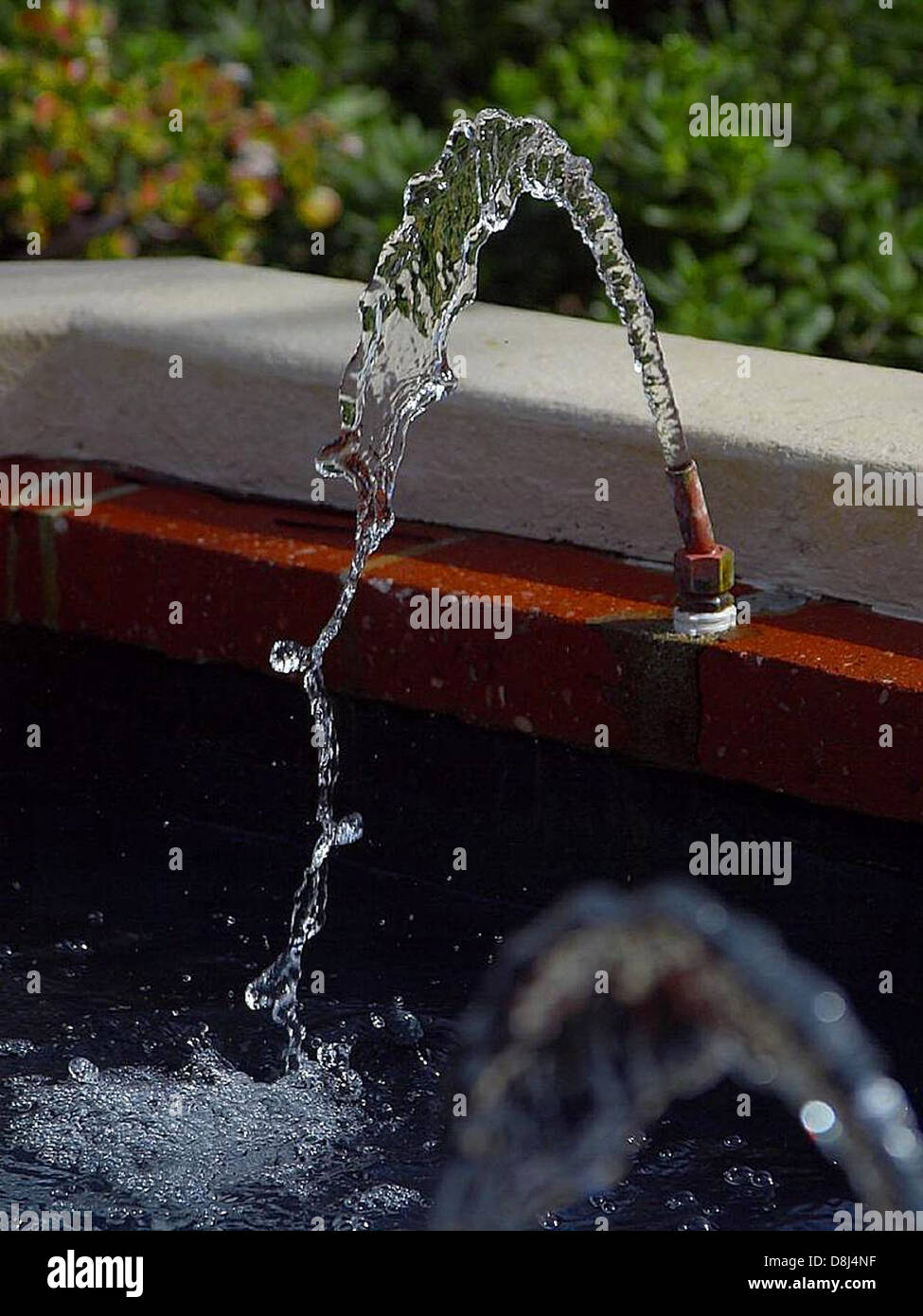 Water spouts from a fountain, creating a dynamic and peaceful display. The water arcs gracefully into the air, sparkling in sunlight, while the surrounding area is calm and serene. Stock Photo