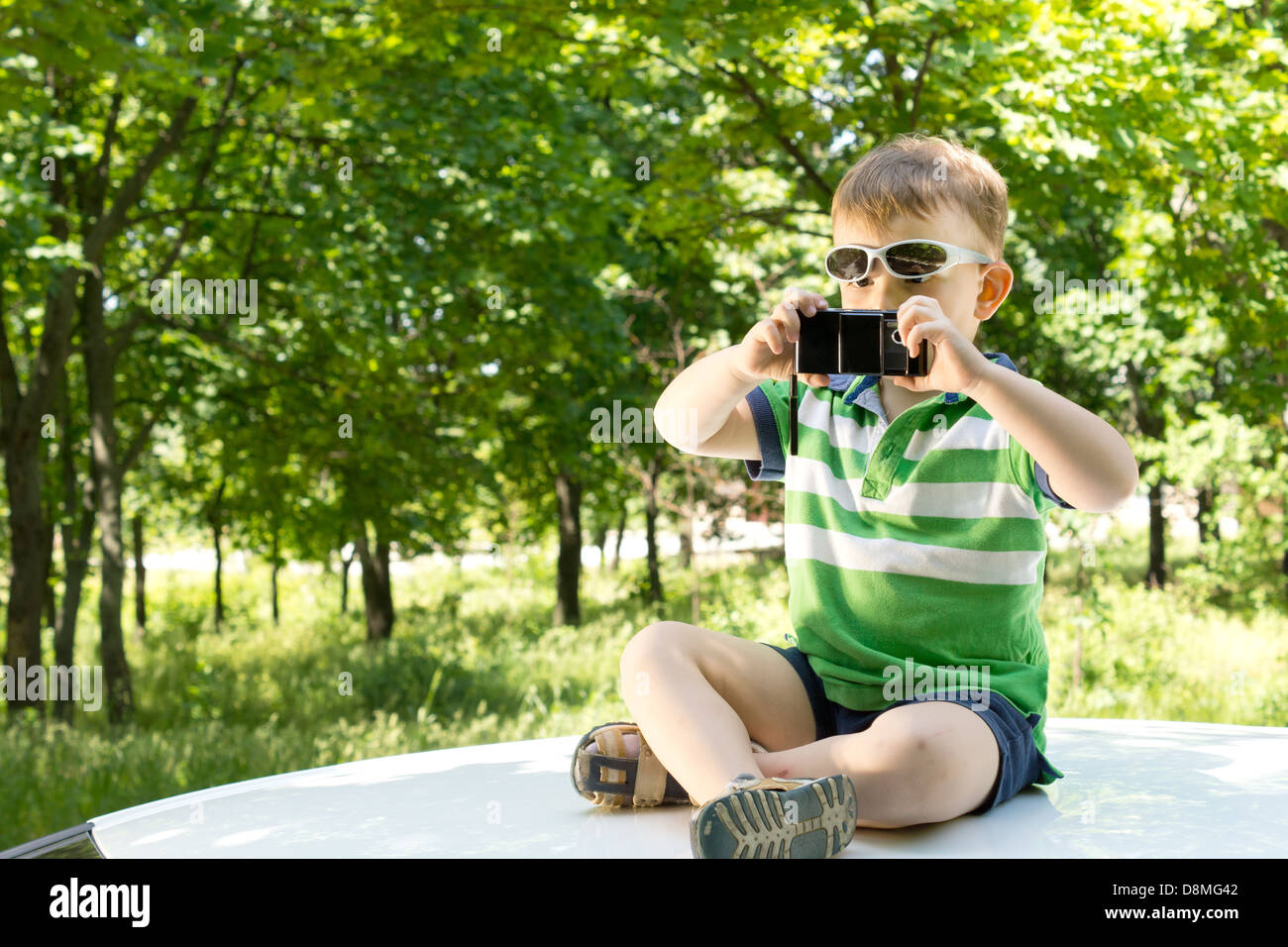 Young blond boy sitting on the top of a white car with crossed legs taking photographs with a mobile phone Stock Photo