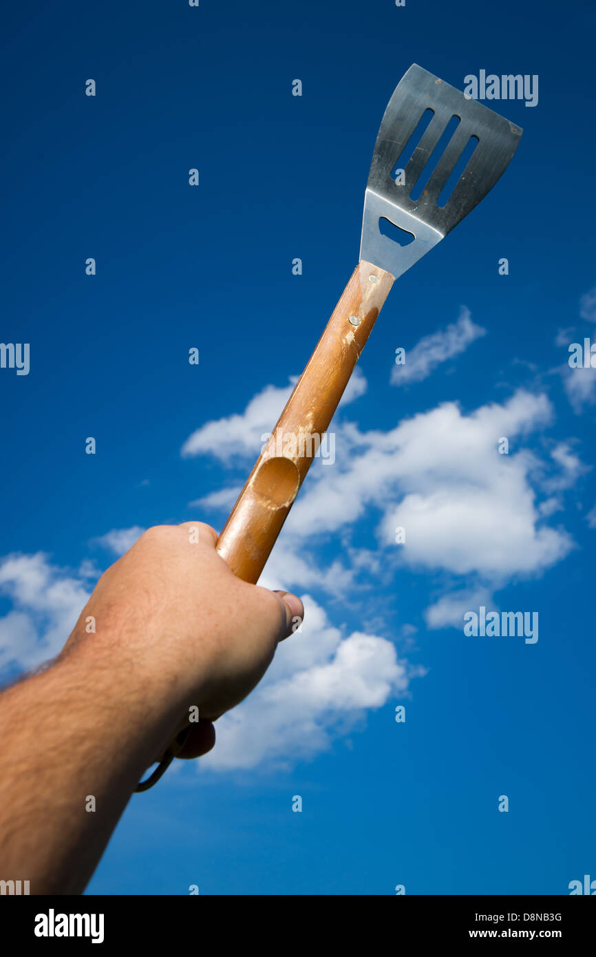 A male hand holds a BBQ spatula aloft against a blue sky with fluffy clouds. Stock Photo