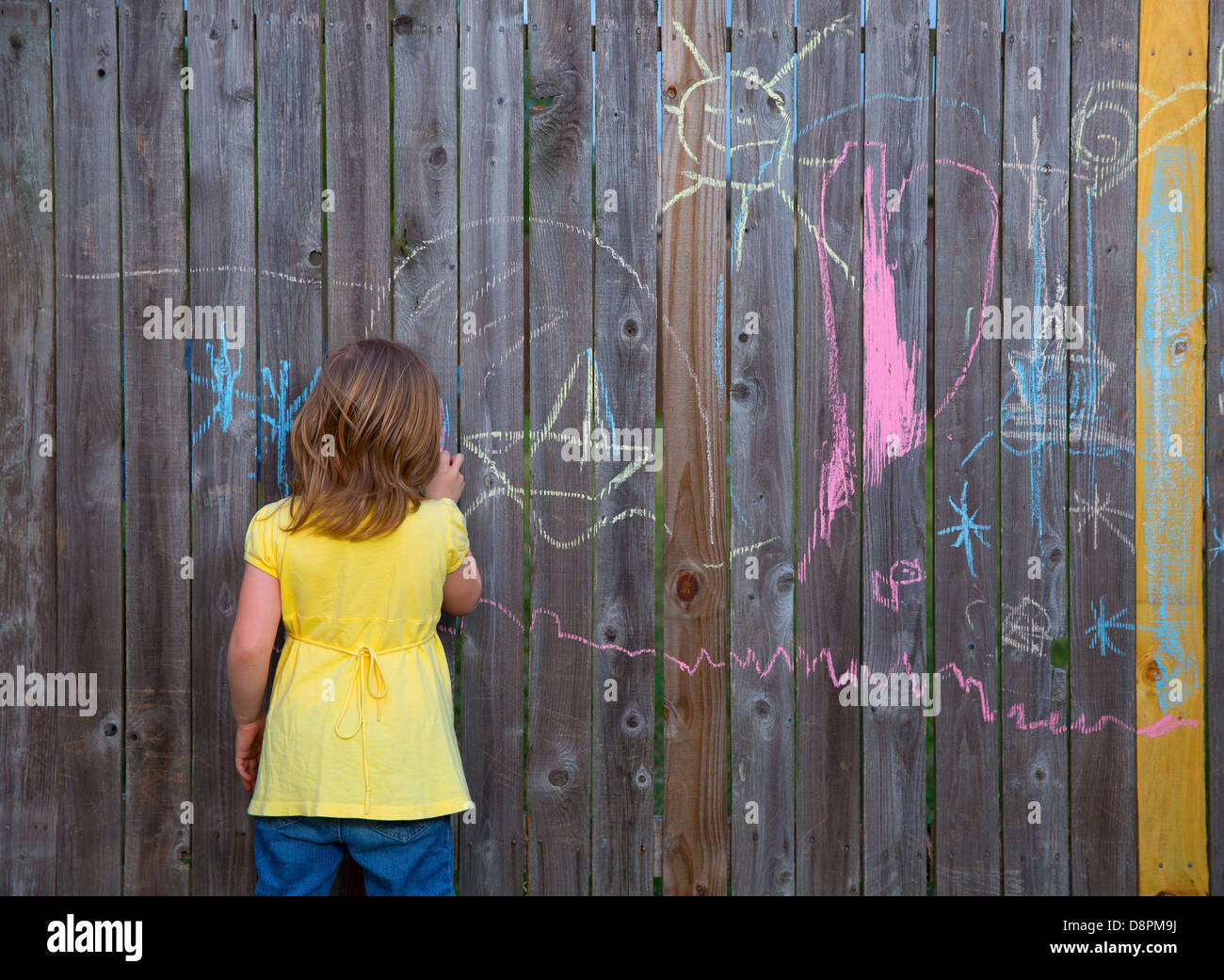 Blonk kid girl playing with drawing chalks in the backyard wooden fence Stock Photo