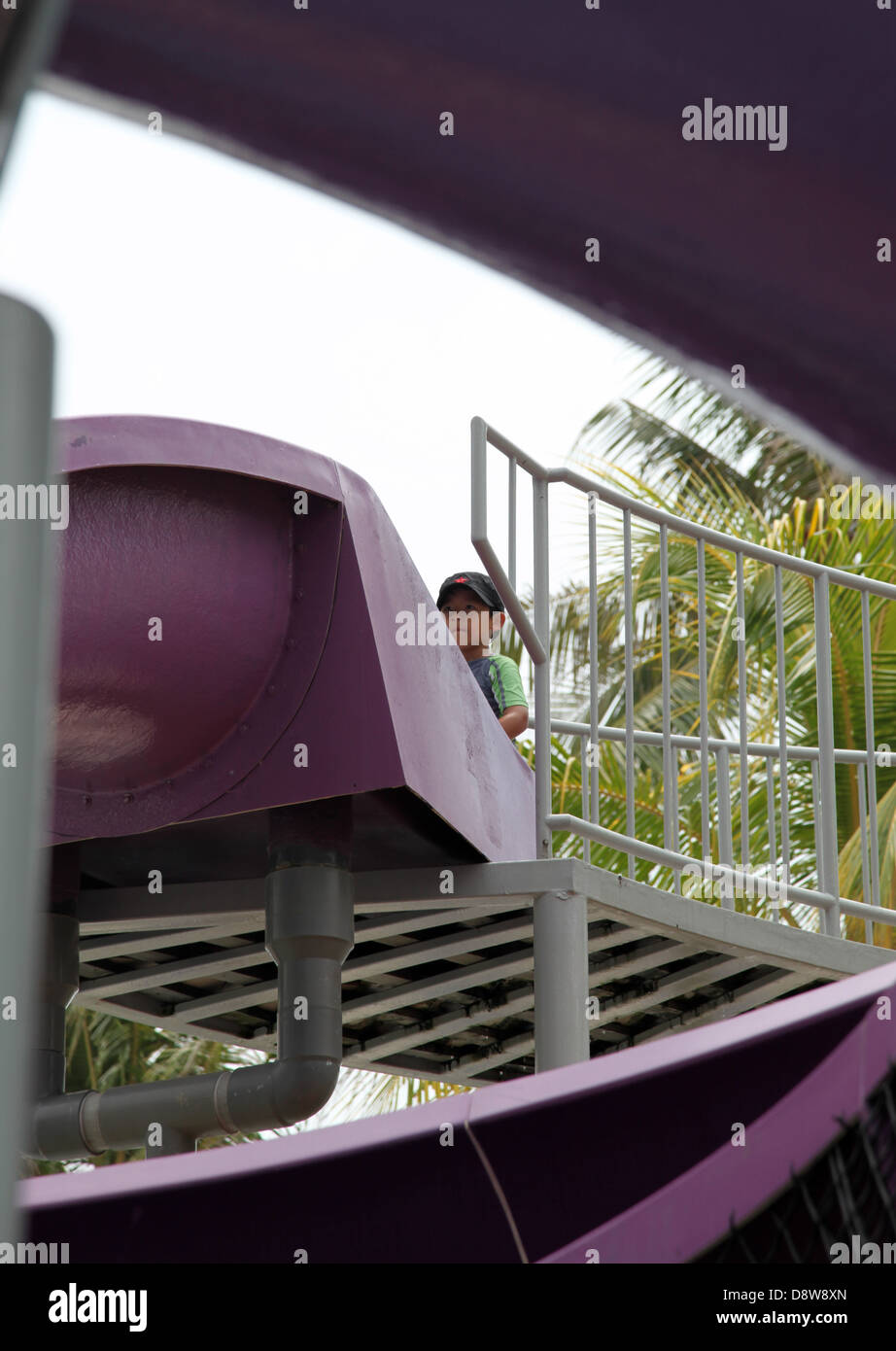 It's a photo of a purple plastic giant slide in a hotel resort. We can see a kid boy who slides down it Stock Photo