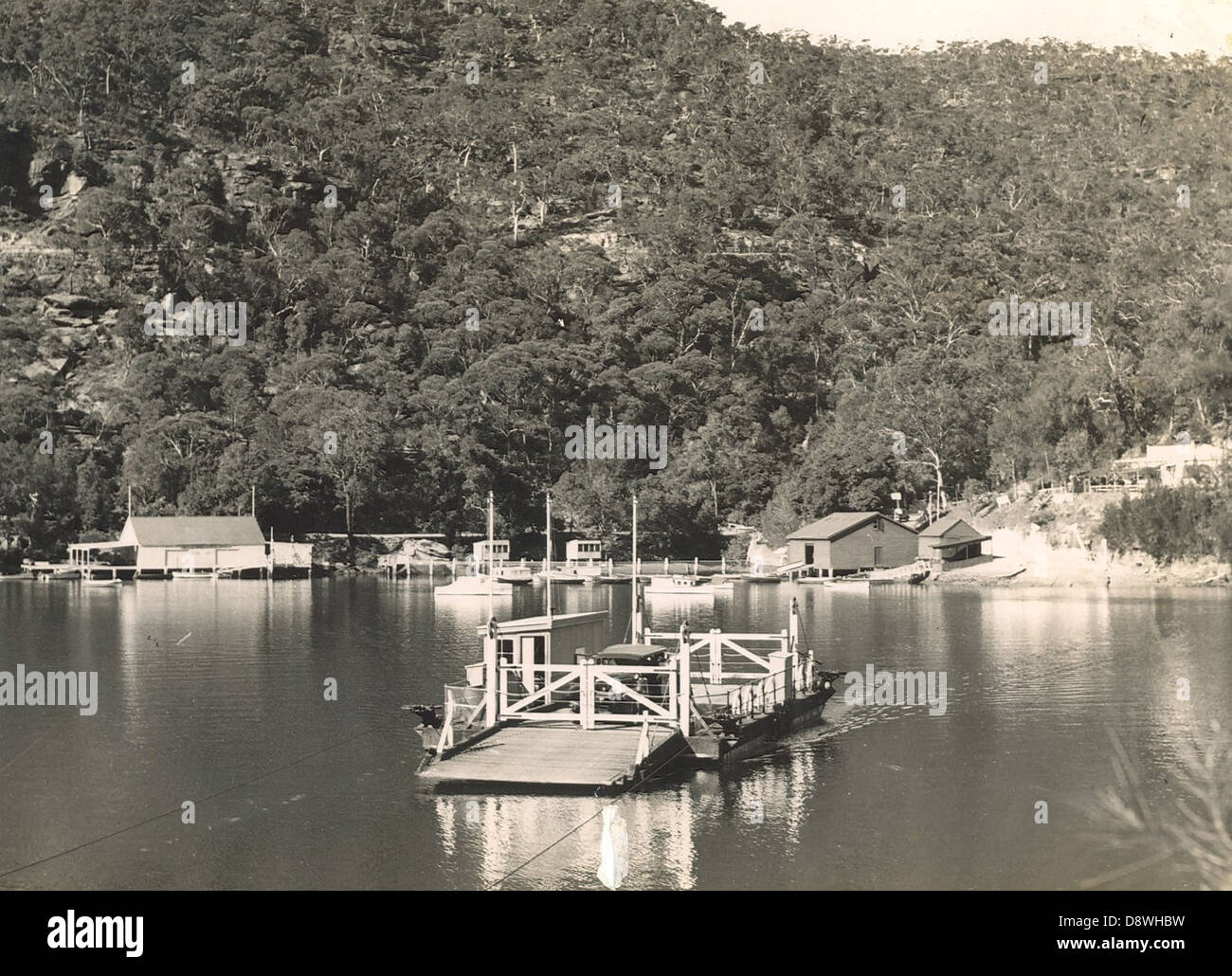 This black-and-white photograph captures a scenic view of Berowra Waters, located on the Hawkesbury River in New South Wales. The image features a boatshed and a car ferry, illustrating the region's transportation and waterfront life in the early 20th century. Stock Photo