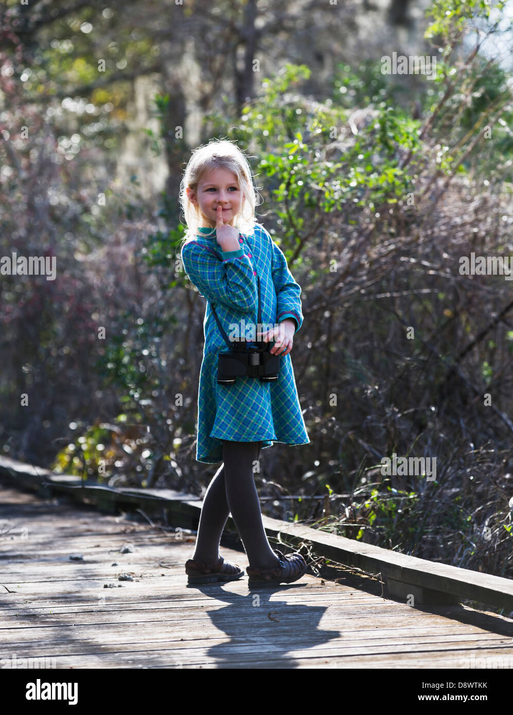 Girl standing on a boardwalk, with her finger to her lips-thinking.Wearing binoculards around her neck. Stock Photo