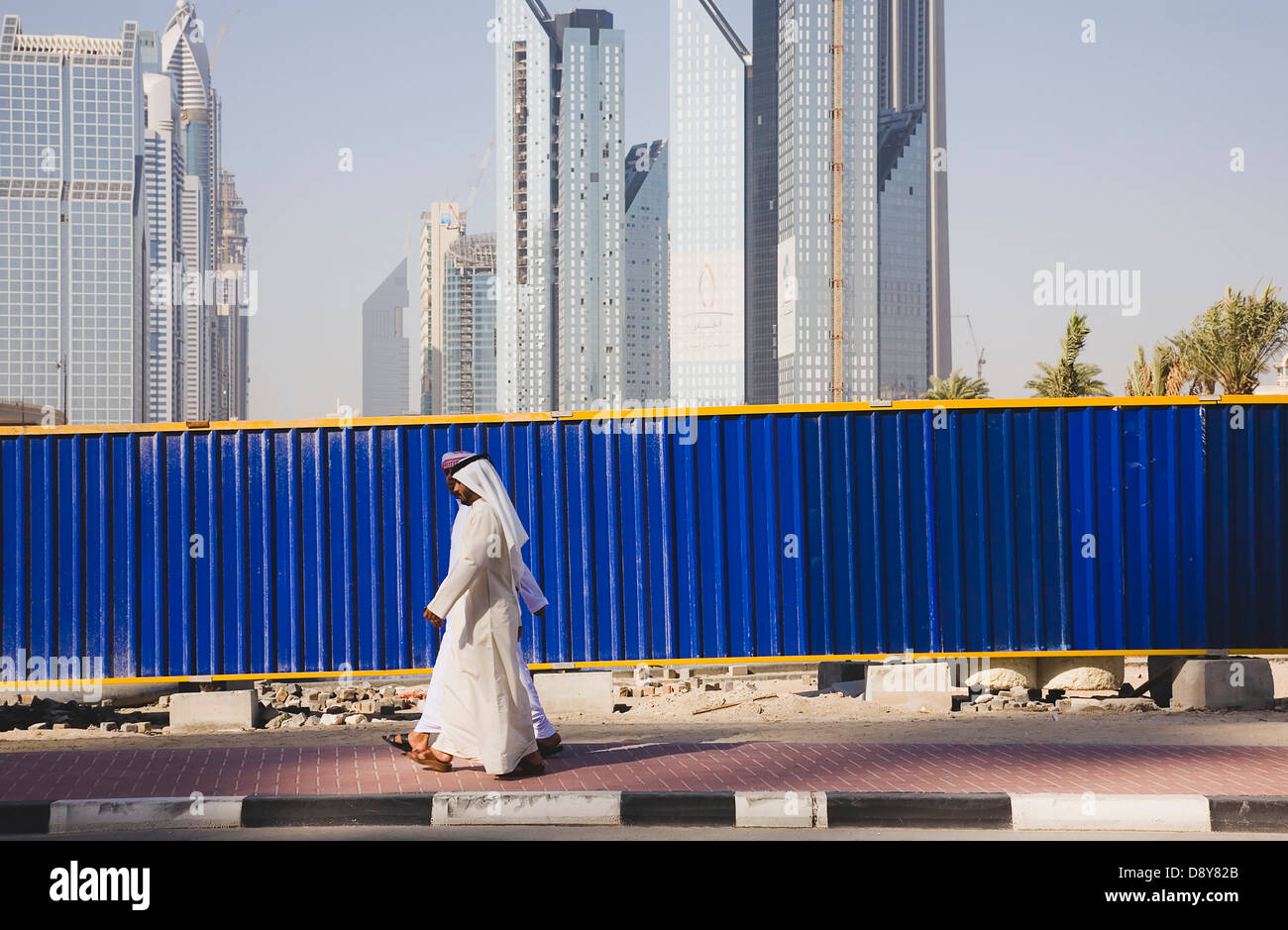 UAE, Dubai, Arab men dressed in traditional Dishdasha walking past construction site with Sheikh Zayed Road behind Stock Photo