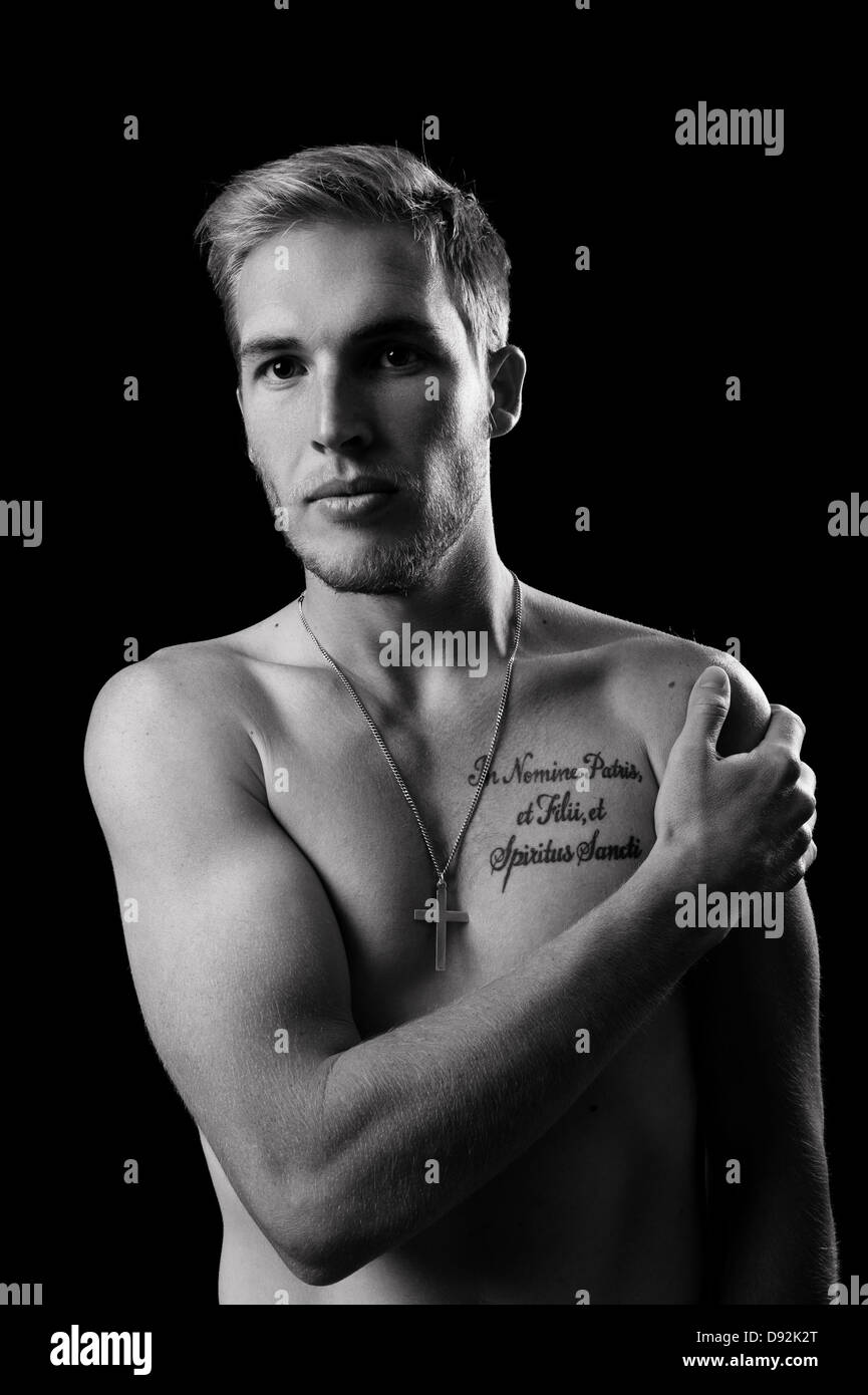 Sexy topless male model with one arm across chest, hand on shoulder, tattoo on chest & cross necklace, low key portrait, strong. Stock Photo