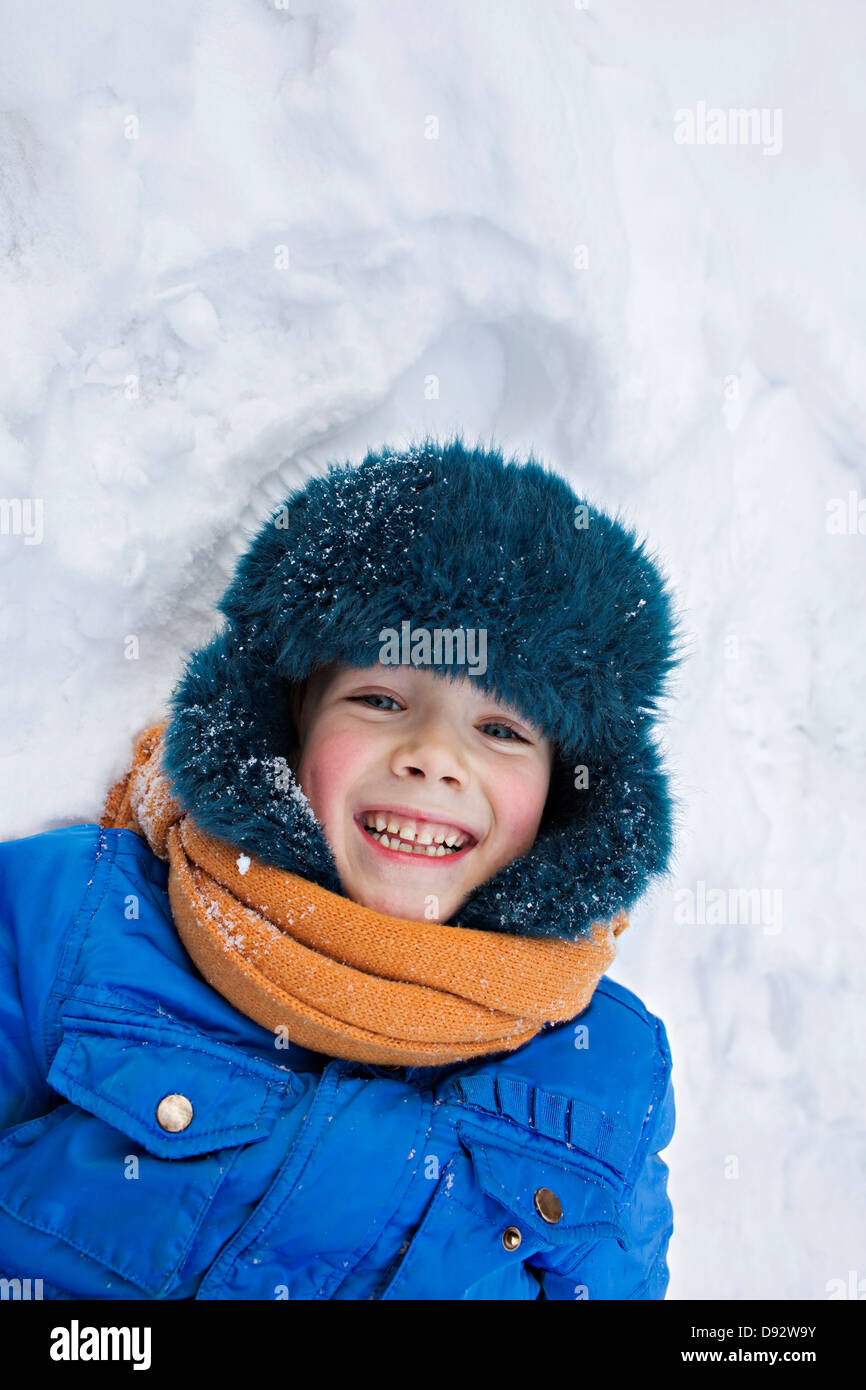 A young cheerful boy wearing warm clothing outdoors lying in the snow Stock Photo