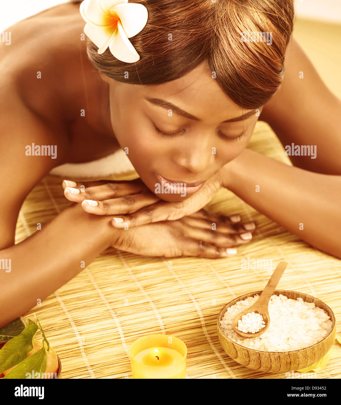 Beautiful african woman with closed eyes and frangipani flower lying down on massage table, enjoying dayspa, luxury spa resort Stock Photo