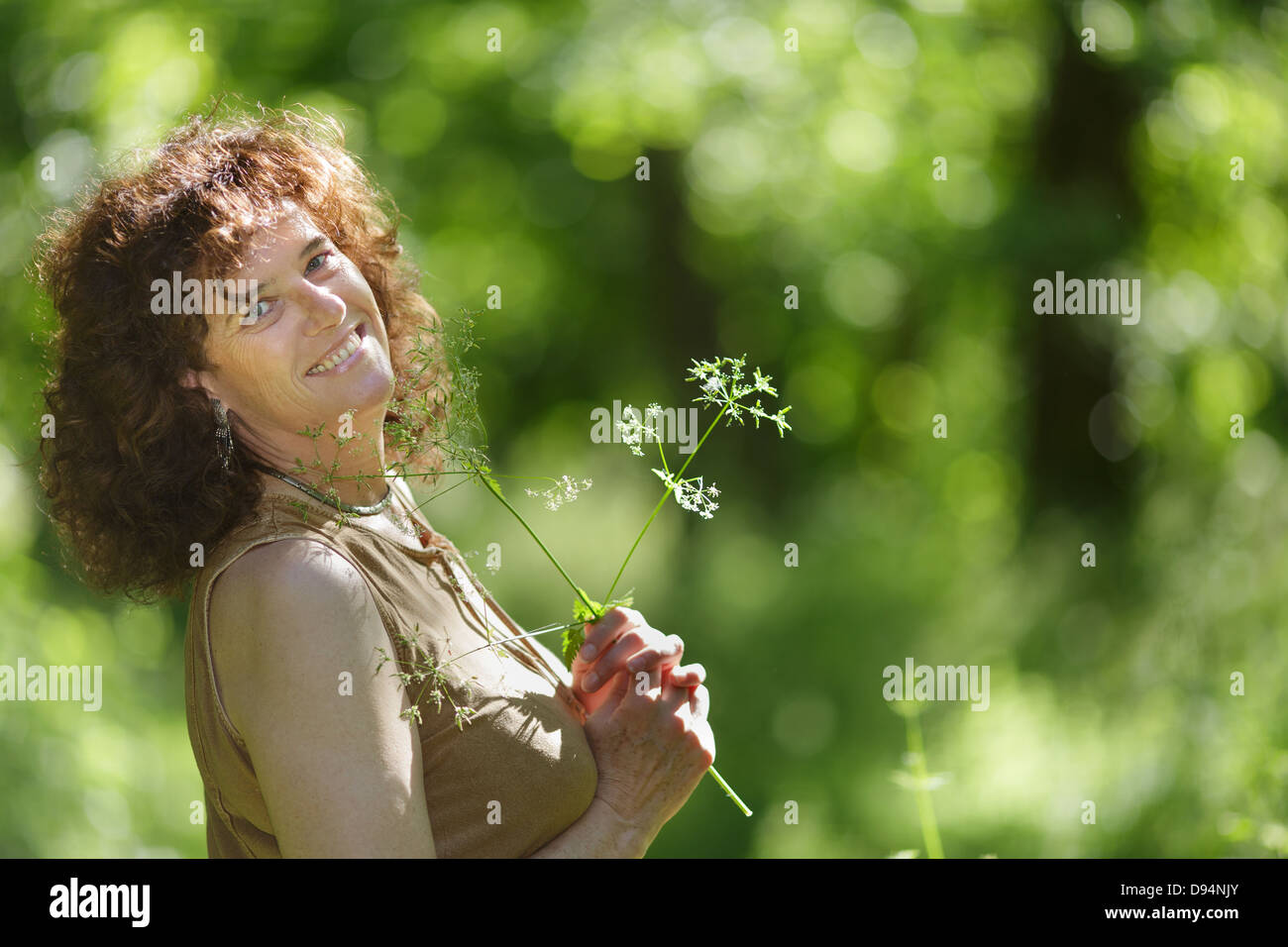 Middle aged mature woman enjoying nature holding wildflower in forest Stock Photo
