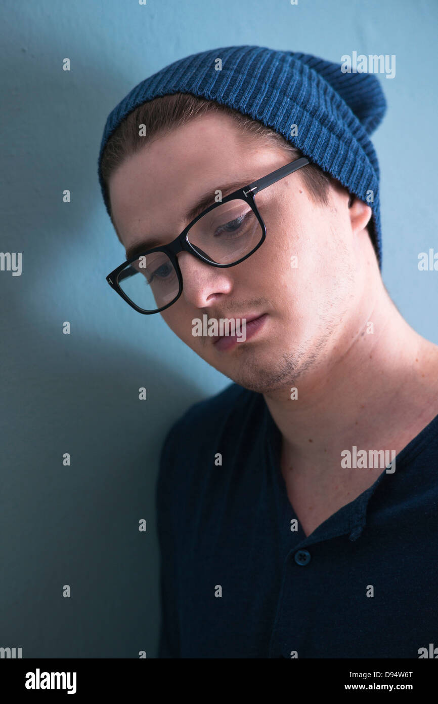 Close-up Portrait of Young Man wearing Woolen Hat and Horn-rimmed Eyeglasses, Looking Downward, Studio Shot on Blue Background Stock Photo
