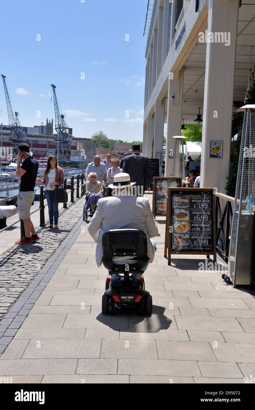 Older man driving electric mobility scooter along Bristol harbourside waterfront walkway, UK Stock Photo