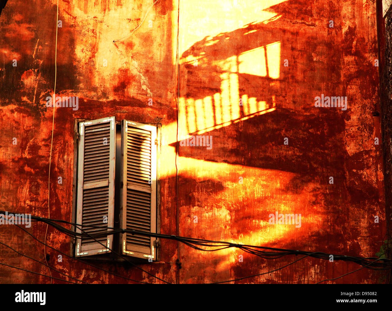 half closed shutters on window and balcony shadow on bright lively colored patchy red and yellow wall Stock Photo