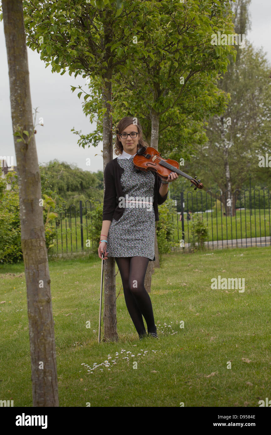 Older teenaged girl holding a violin standing in a lawn with trees. Stock Photo