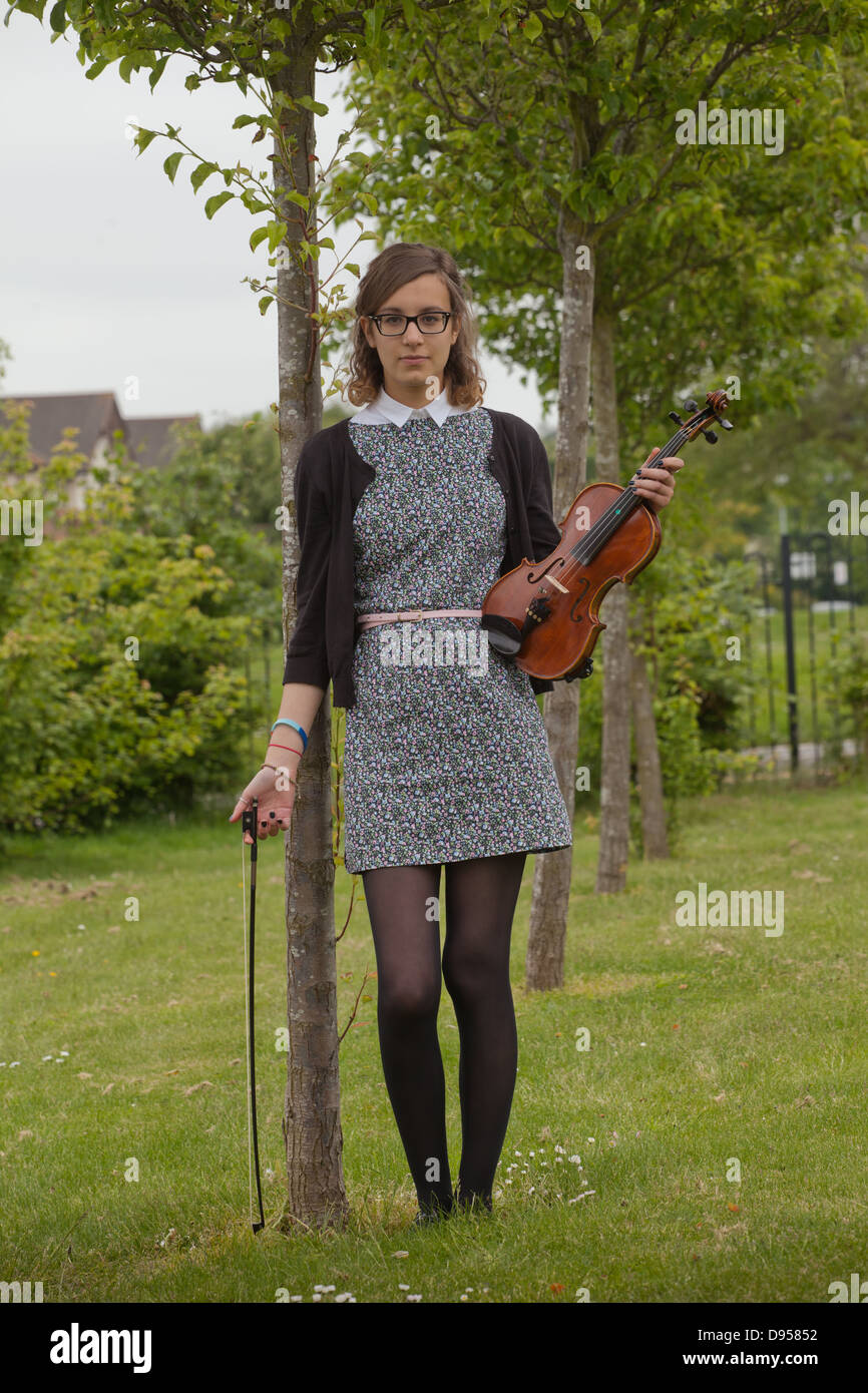 Older teenaged girl holding a violin standing in a lawn with trees. Stock Photo