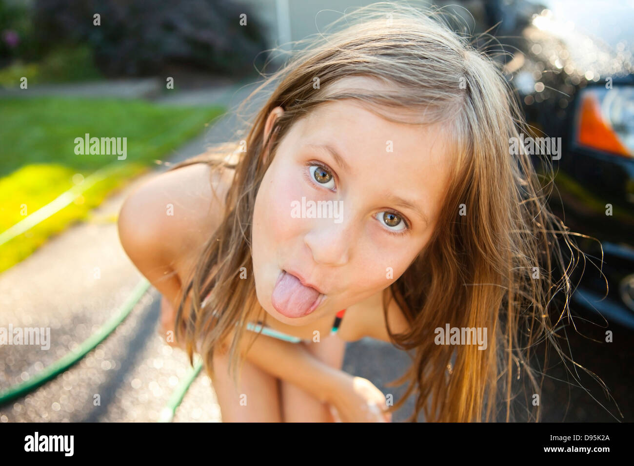 Portrait young girl sticking her tongue out while washing car in driveway their home on sunny summer afternoon in Portland Stock Photo