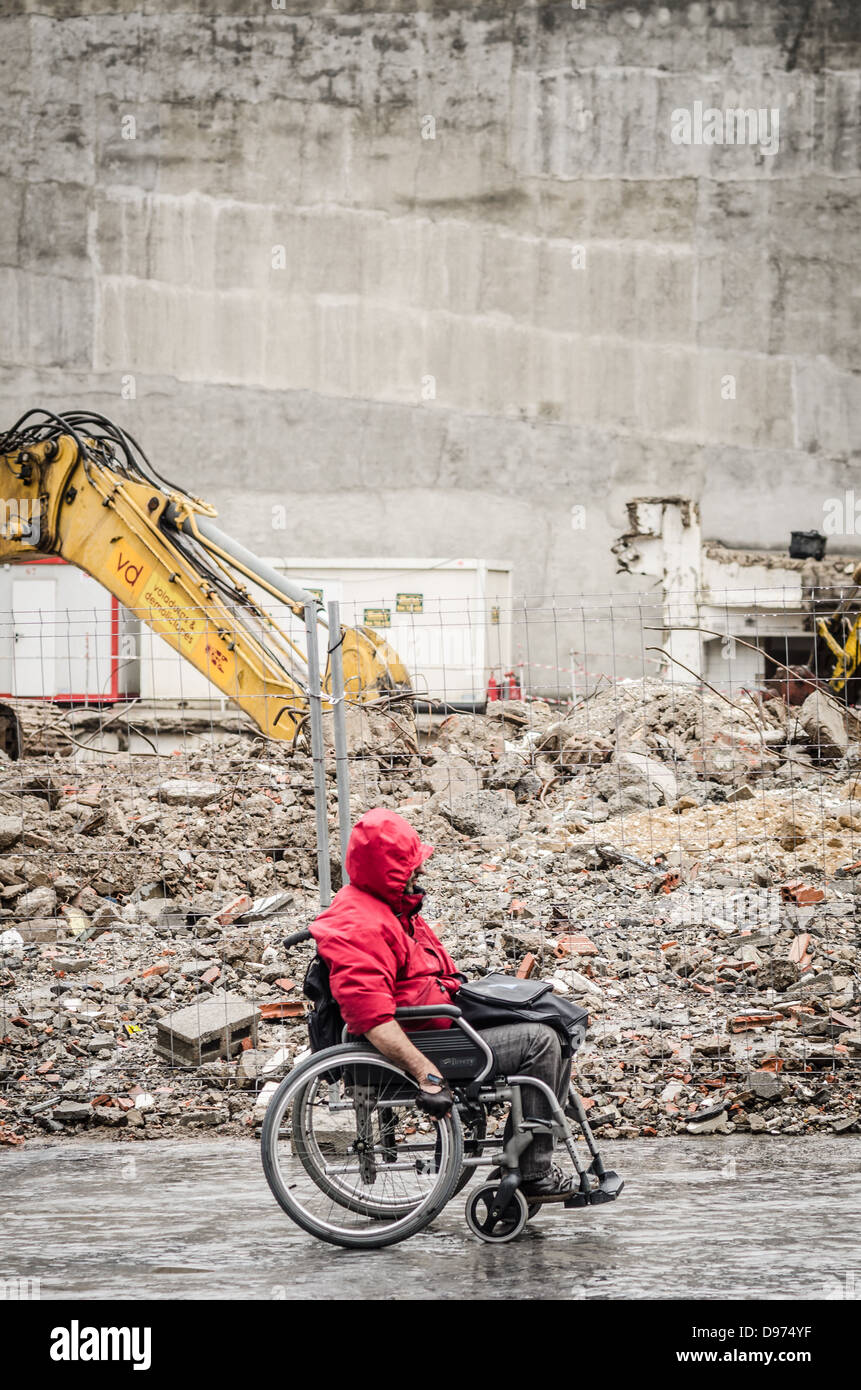 A man in a wheelchair struggles in bad weather along side a building site. Stock Photo