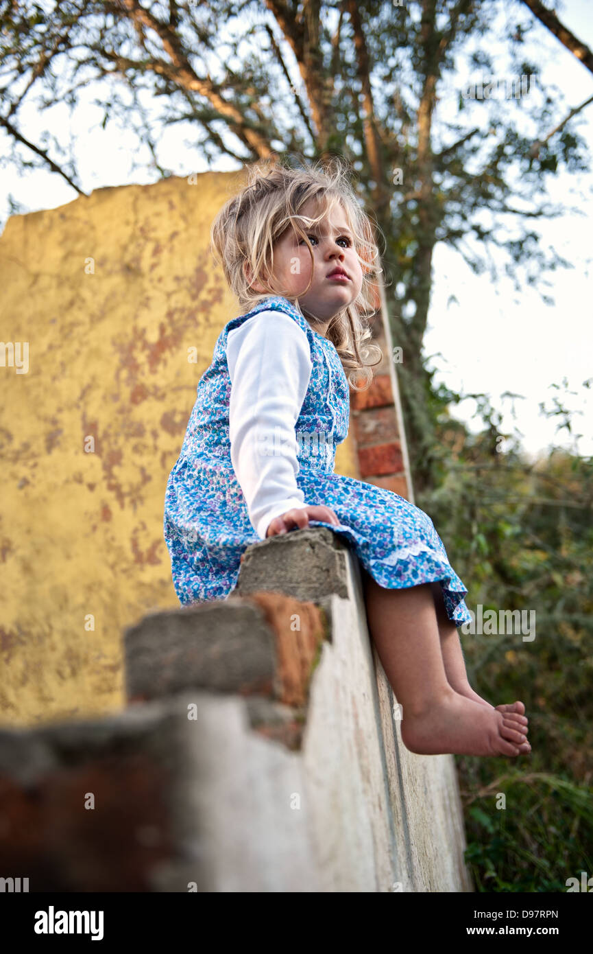 Blonde toddler with blue flowery dress and barefoot, sitting on an old dilapidated brick wall staring up emotionless. Stock Photo