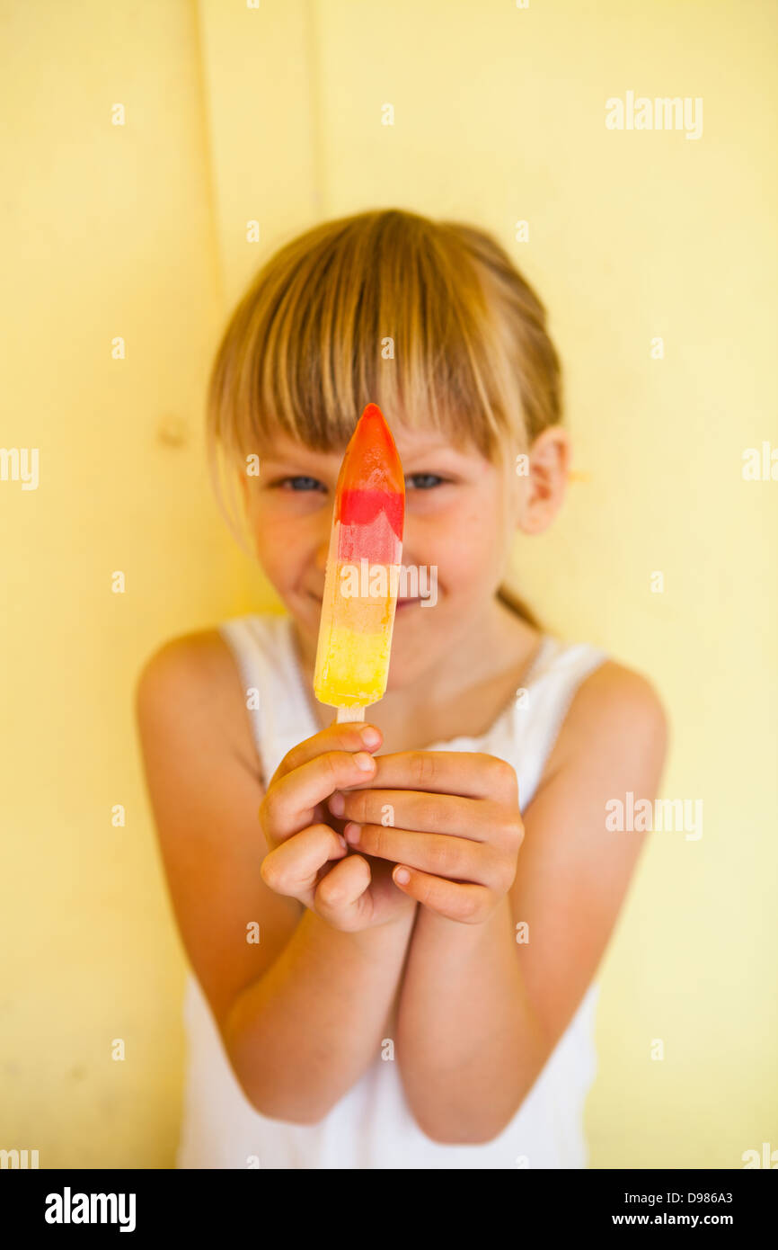 Smiling young girl holding up multicolored popsicle against yellow colored background Stock Photo