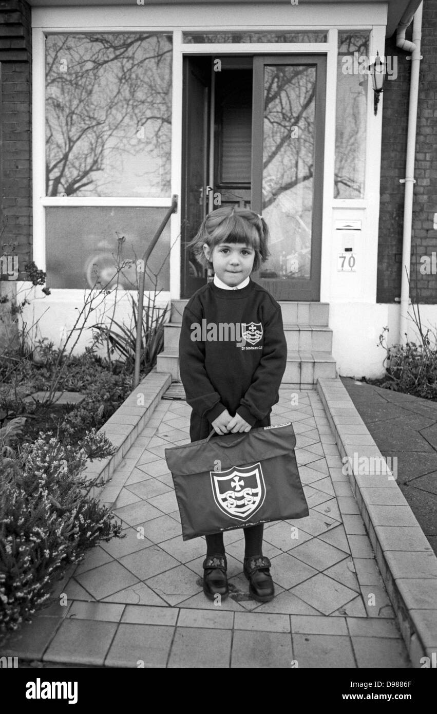 A 5 year-old girl stands outside her south London home on the first day of proper school, a momentous day and a rite of passage. Standing on the path by the front door of an Edwardian period south London home, the girl holds a brand new book bag with the initials of her local school of St Saviour's, repeated on her school jumper. She looks calm but is inwardly nervous of the day about to unfold - a rite of passage for every schoolchild, climbing the ladder of life. Stock Photo