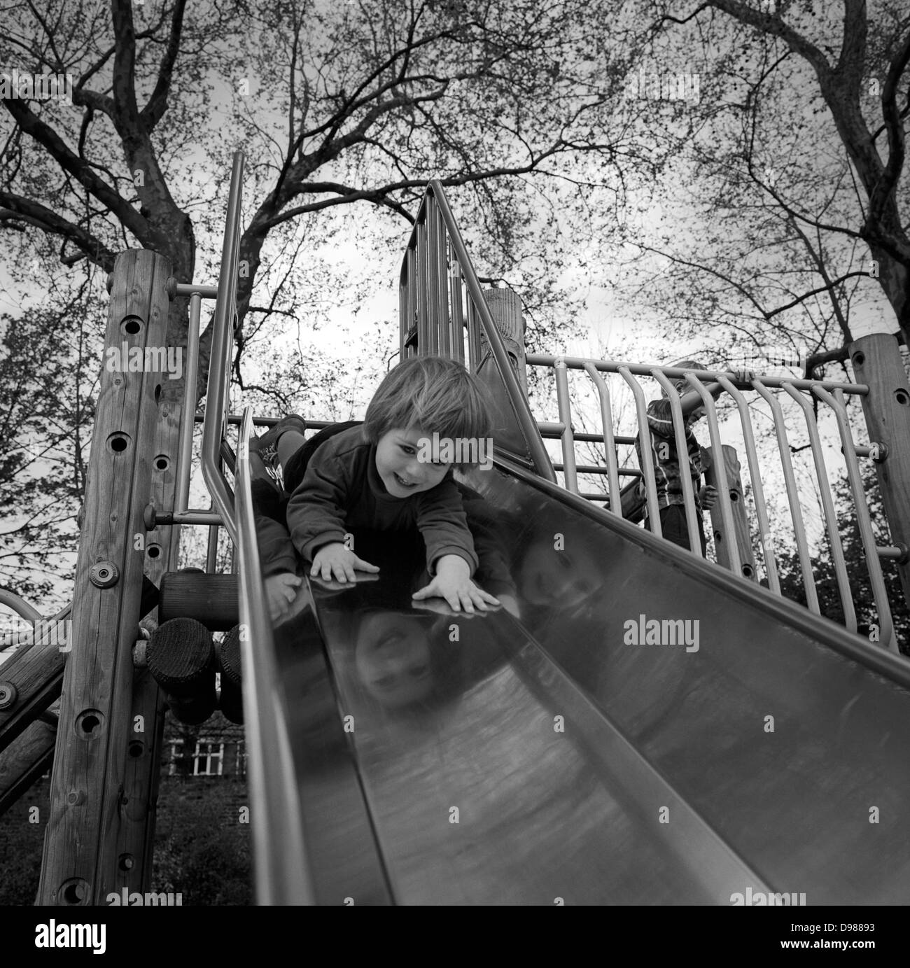 With hands outstretched and a nervous smile on her face, a 3 year-old goes head first down a slide in her local park in south London. The gradient helps the girl on her downward journey as the tentatively slides down with her fingers feeling the polished surface, her smiling, confident face reflected on the three sides of the slide's metal. Tall London plane trees rise above in this public London space. Stock Photo