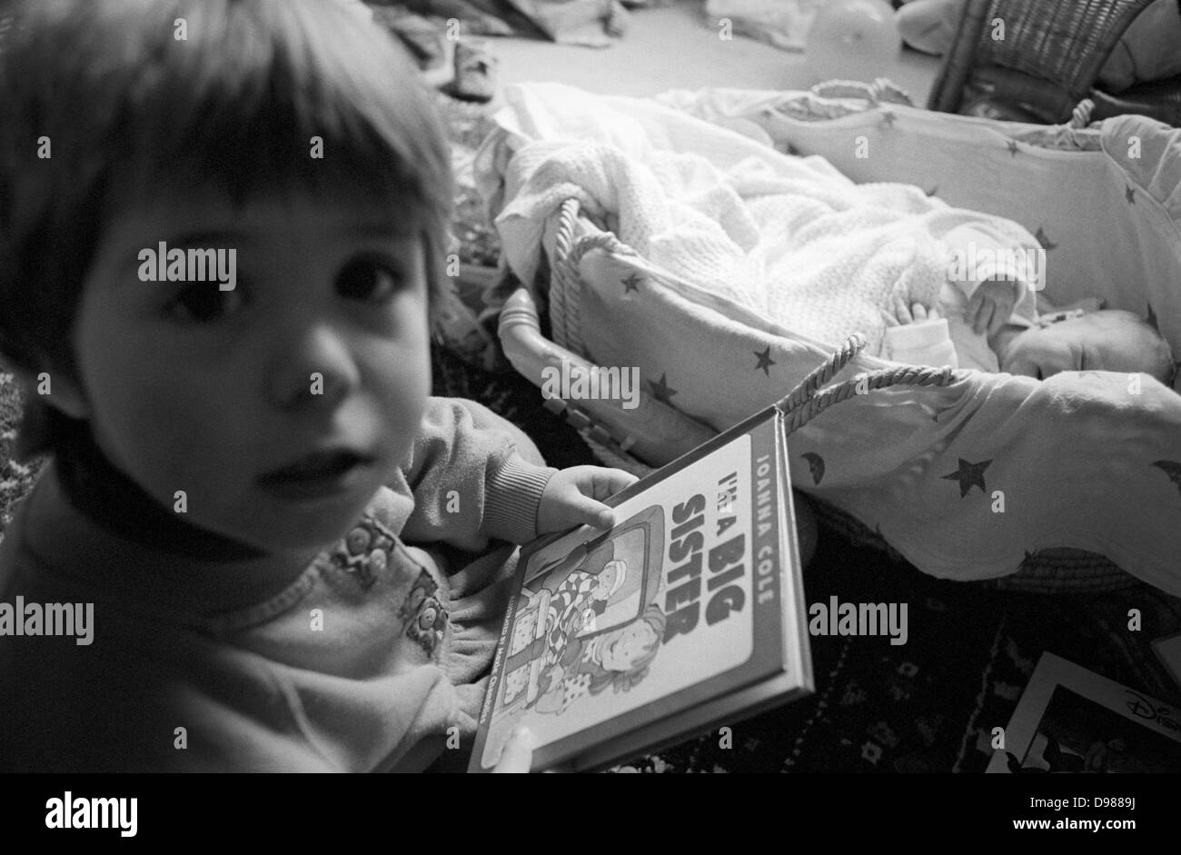 A 2 year-old girl reads a book about being a big sister, coming to terms with role-play, the idea of having a newborn baby brother in his basket at home in south London. The siblings are in their south London home, the girl sitting up on the carpet and her brother - only a few weeks old - is starting to wake from a mid-morning sleep. The book being read is 'I'm a big sister' by writer Joanna Cole, written to help young girl's overcome the shock of suddenly no longer being the most important member of a young family, helping her deal with a new status as the eldest but more mature child. Stock Photo