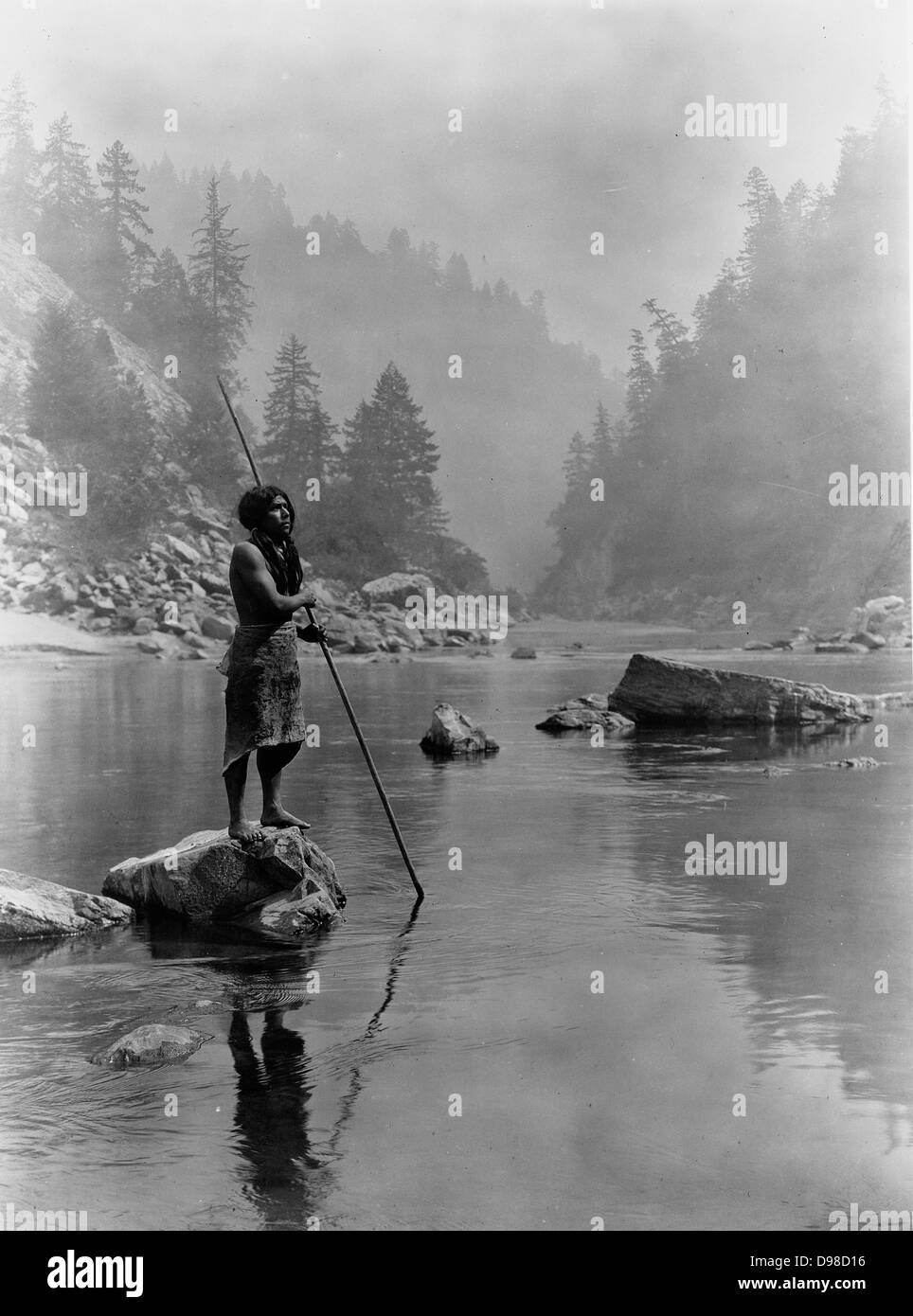 Hupa man with spear, standing on rock midstream, in background, fog partially obscures trees on mountainsides, 1923. Photograph by Edward Curtis (1868-1952). Stock Photo