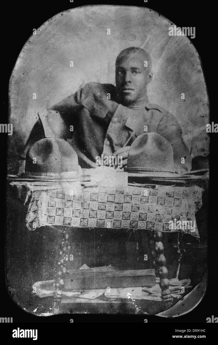 World War I 1914-1918: African-American soldier seated behind table, pencil in hand and facing front, with two hats on table. Stock Photo