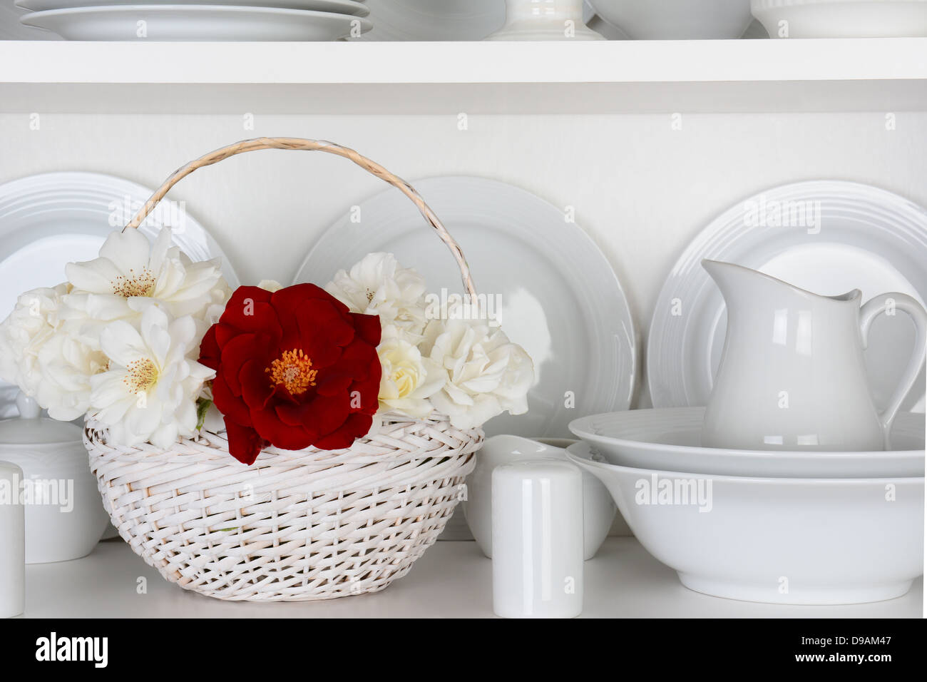 Closeup of a basket of roses on the shelf of a cupboard full of white plates. Items include, plates, saucers, bowls Stock Photo