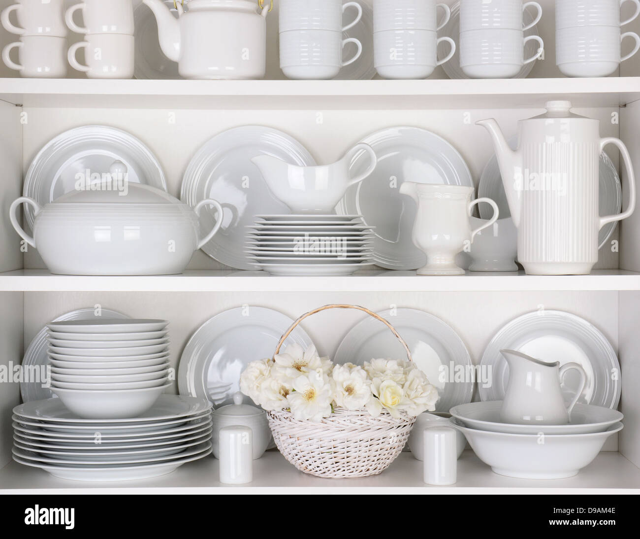 Closeup of white plates and dinnerware in a cupboard. A basket of white roses is centered on the bottom shelf. Stock Photo
