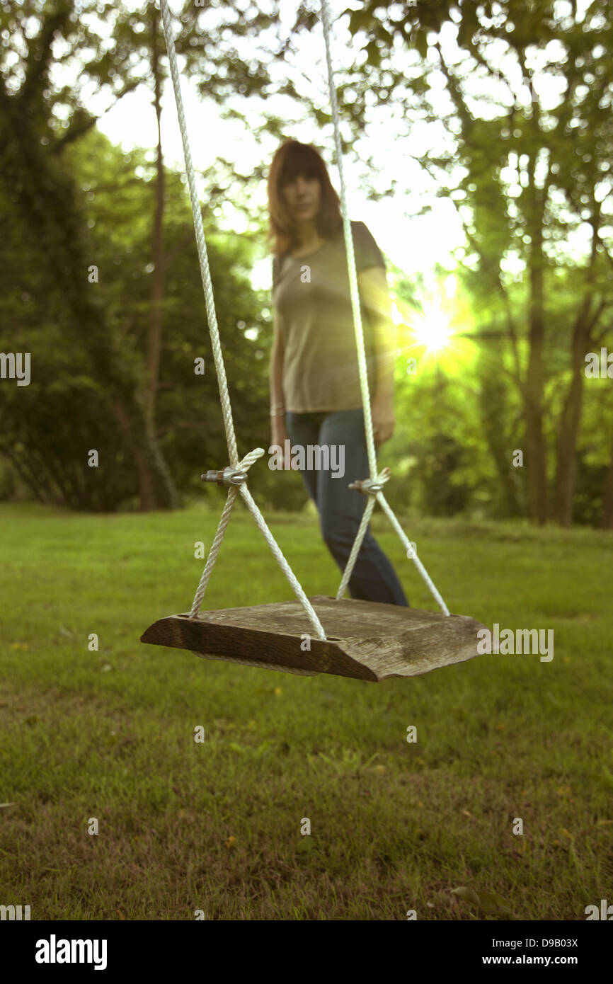 A pensive woman looking at an empty garden swing, the sun shining through the trees. Stock Photo