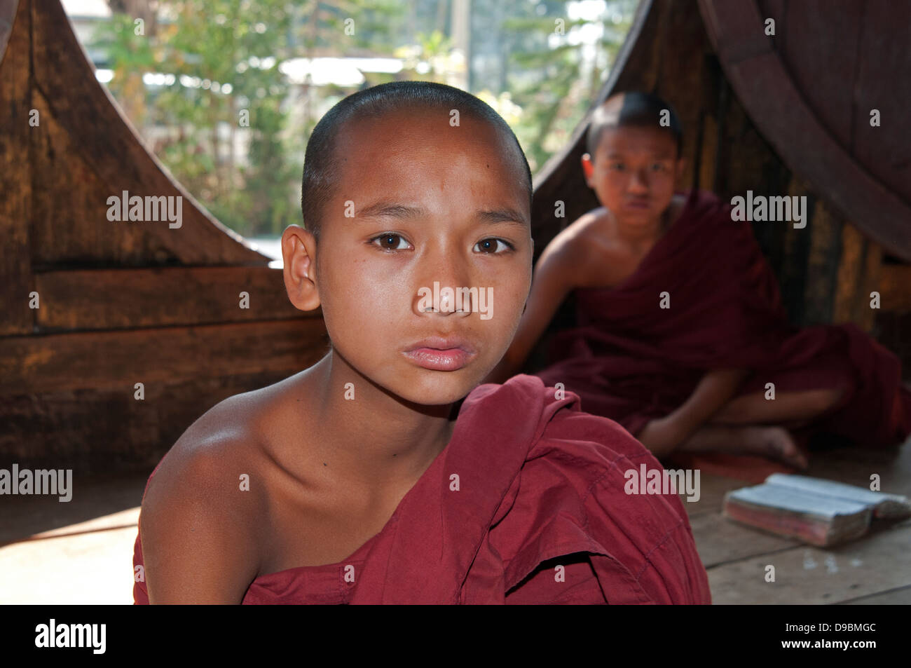 Two novice Buddhist monks in red robes sitting crossed legged in wooden monastery by round window Myanmar Stock Photo