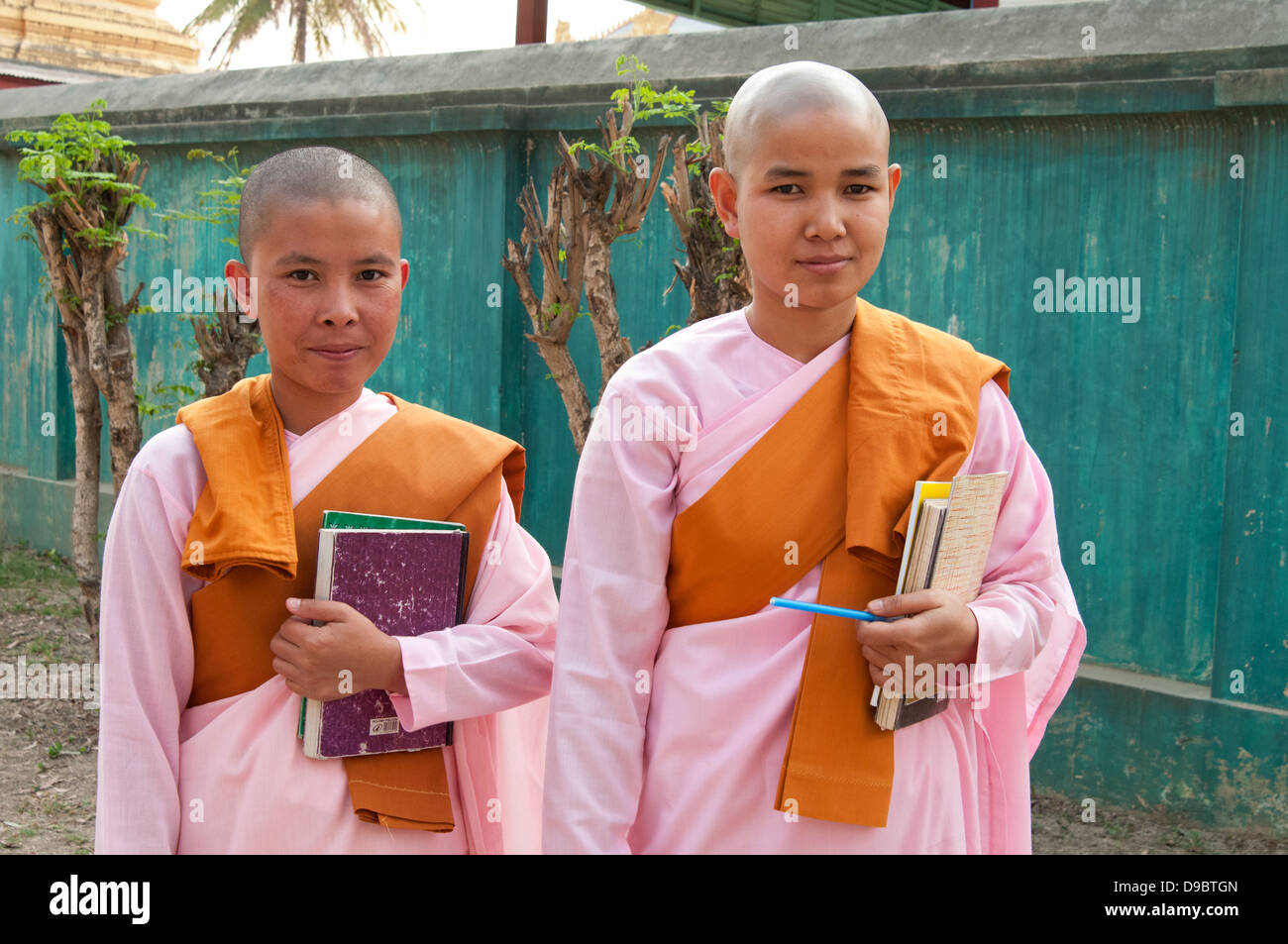 Two Buddhist nuns wearing pink robes carrying books stand in front of a green temple wall street Myanmar (Burma) Stock Photo