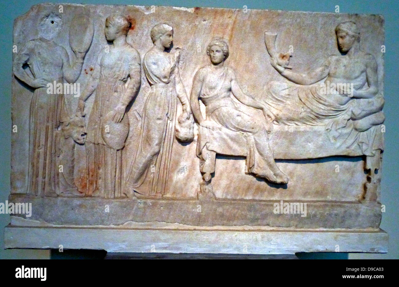 Votive relief.  Marble.  Found in the Piraeus.  Dionysus is shown on a kline, holding a rhyton and a phiale.  At the foot of his kline stands a woman, Paideia, with her face turned towards a man who holds a theatrical mask in his left hand.  Two actors are shown at left.  The relief was dedicated by actors to the god Dionysus after a theatrical performance. circa 400 BC. Stock Photo