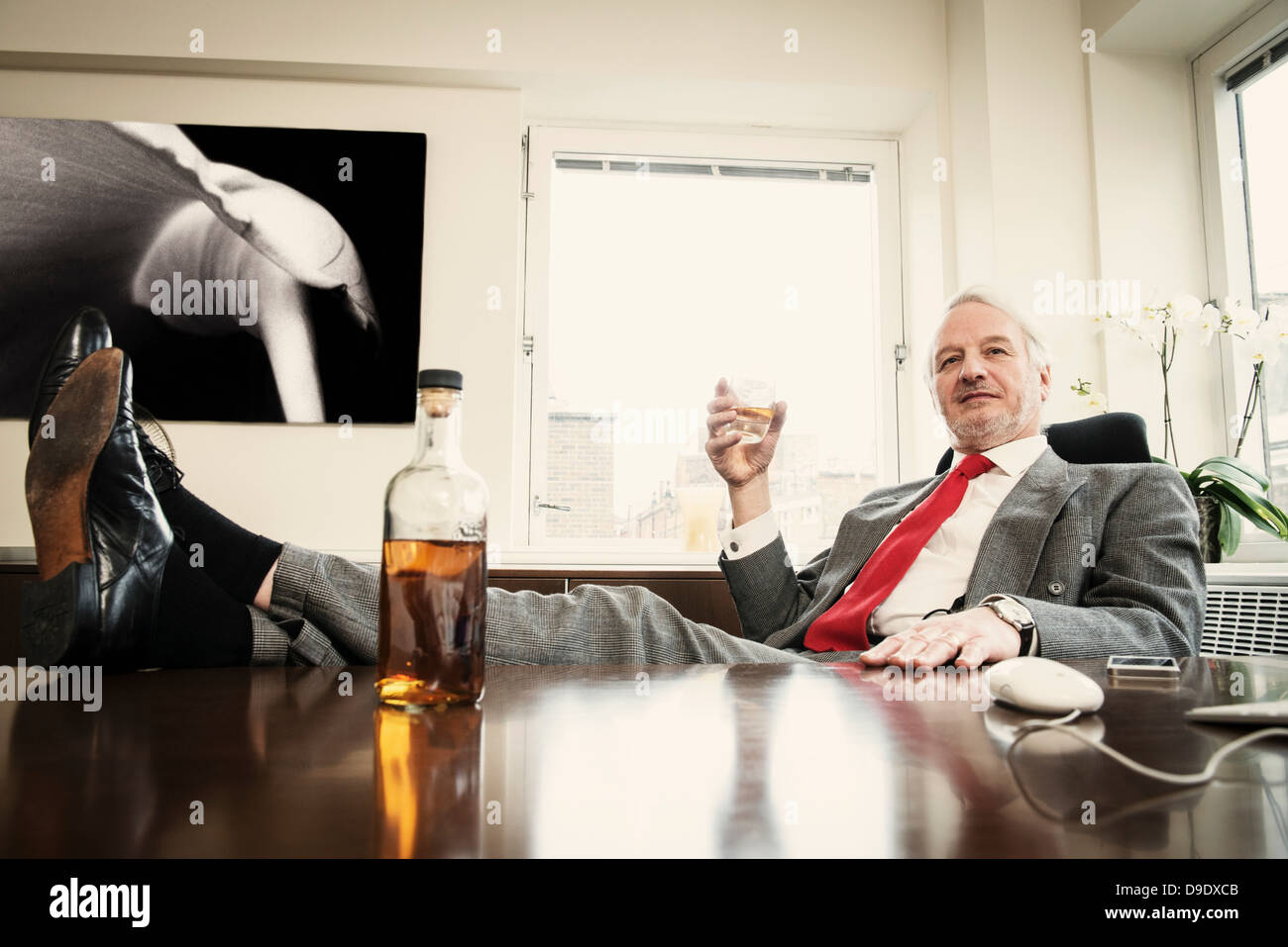 Senior Businessman With Feet Up On Desk Drinking Spirits
