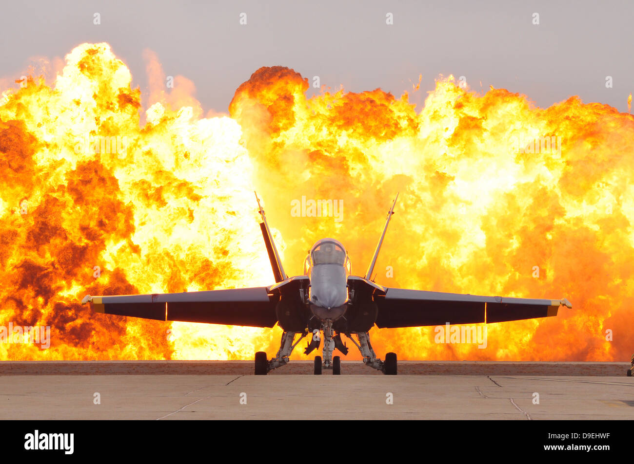 An F/A-18 Hornet sits on the flight line as a wall of fire detonates behind it. Stock Photo