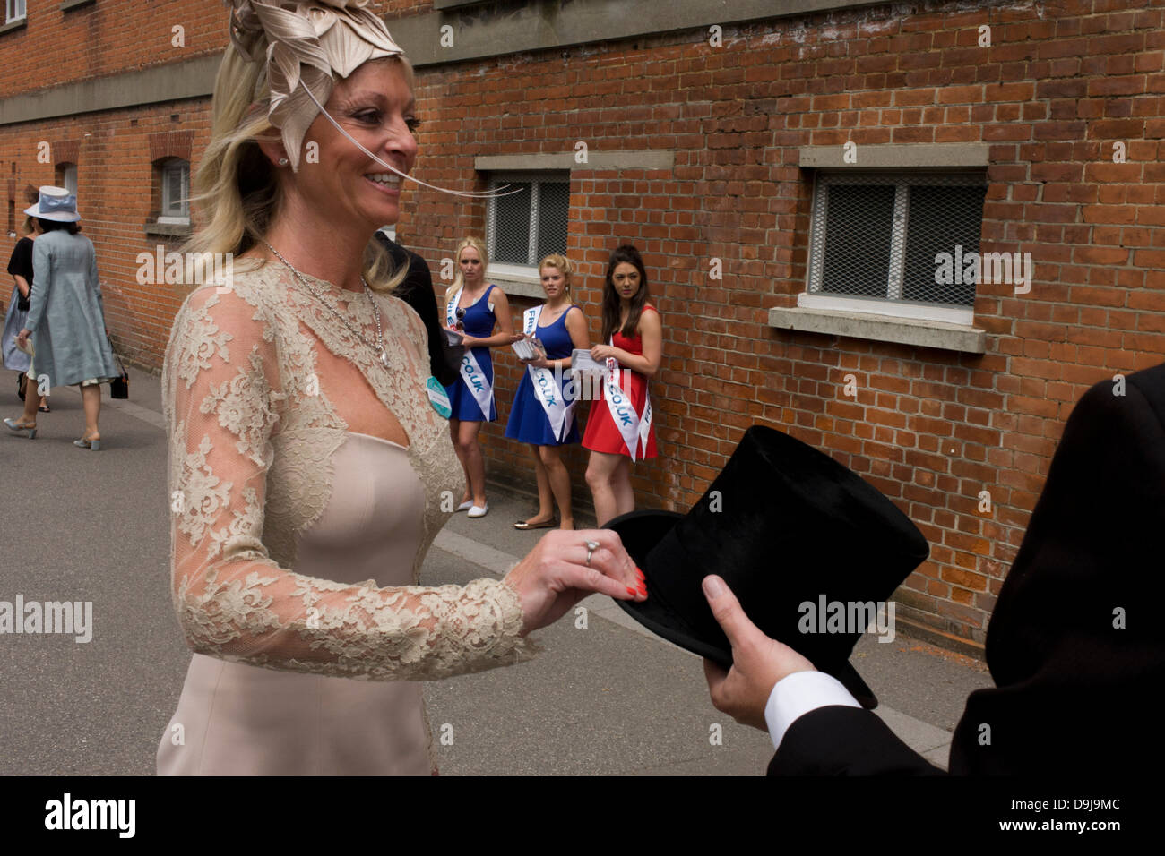 With betting promo girls looking on, a lady partner of an unseen gentleman dressed in formal menswear, passes back his top hat during the annual Royal Ascot horseracing festival in Berkshire, England. Royal Ascot is one of Europe's most famous race meetings, and dates back to 1711. Queen Elizabeth and various members of the British Royal Family attend. Held every June, it's one of the main dates on the English sporting calendar and summer social season. Over 300,000 people make the annual visit to Berkshire during Royal Ascot week, making this Europe’s best-attended race meeting with over £3m Stock Photo