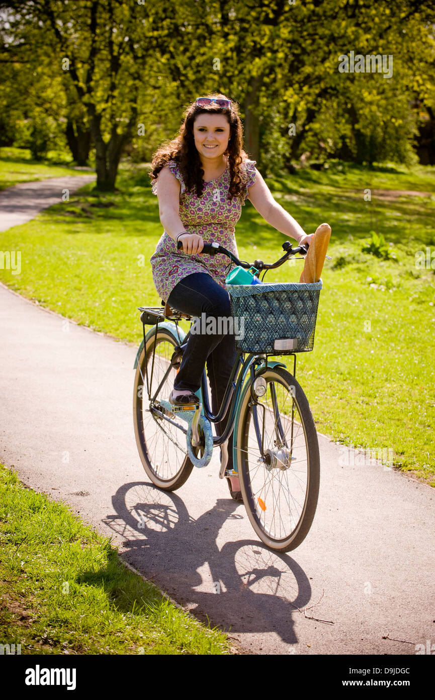Young caucasian woman riding a bike with a flask and a baguette in the basket, in a UK park, in summer Stock Photo