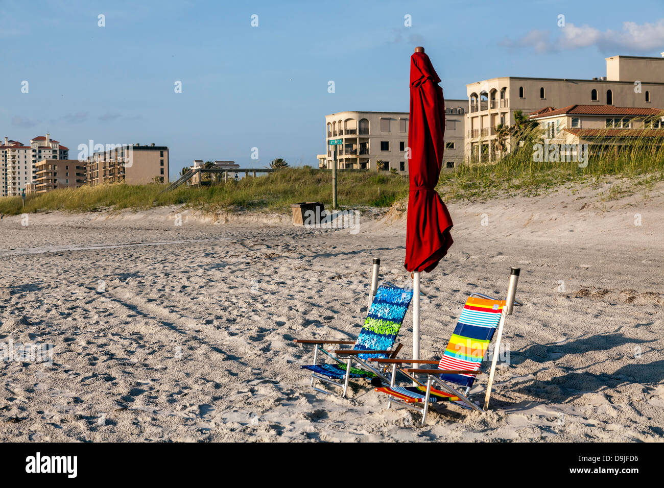 Colorful striped folding beach chairs and folded red umbrella with buildings in the background