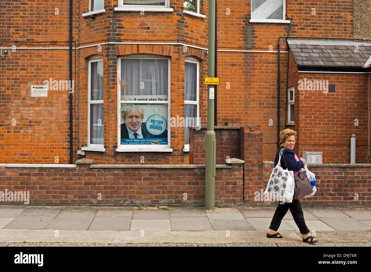 Woman walks past house which has a Boris Johnson Mayoral election campaign poster in the window. She looks towards camera. Stock Photo
