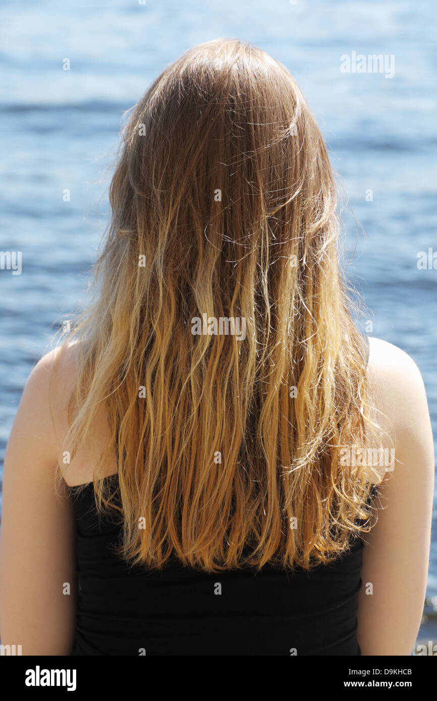 Girl with blond long hair sitting near the water back to the viewer Stock Photo