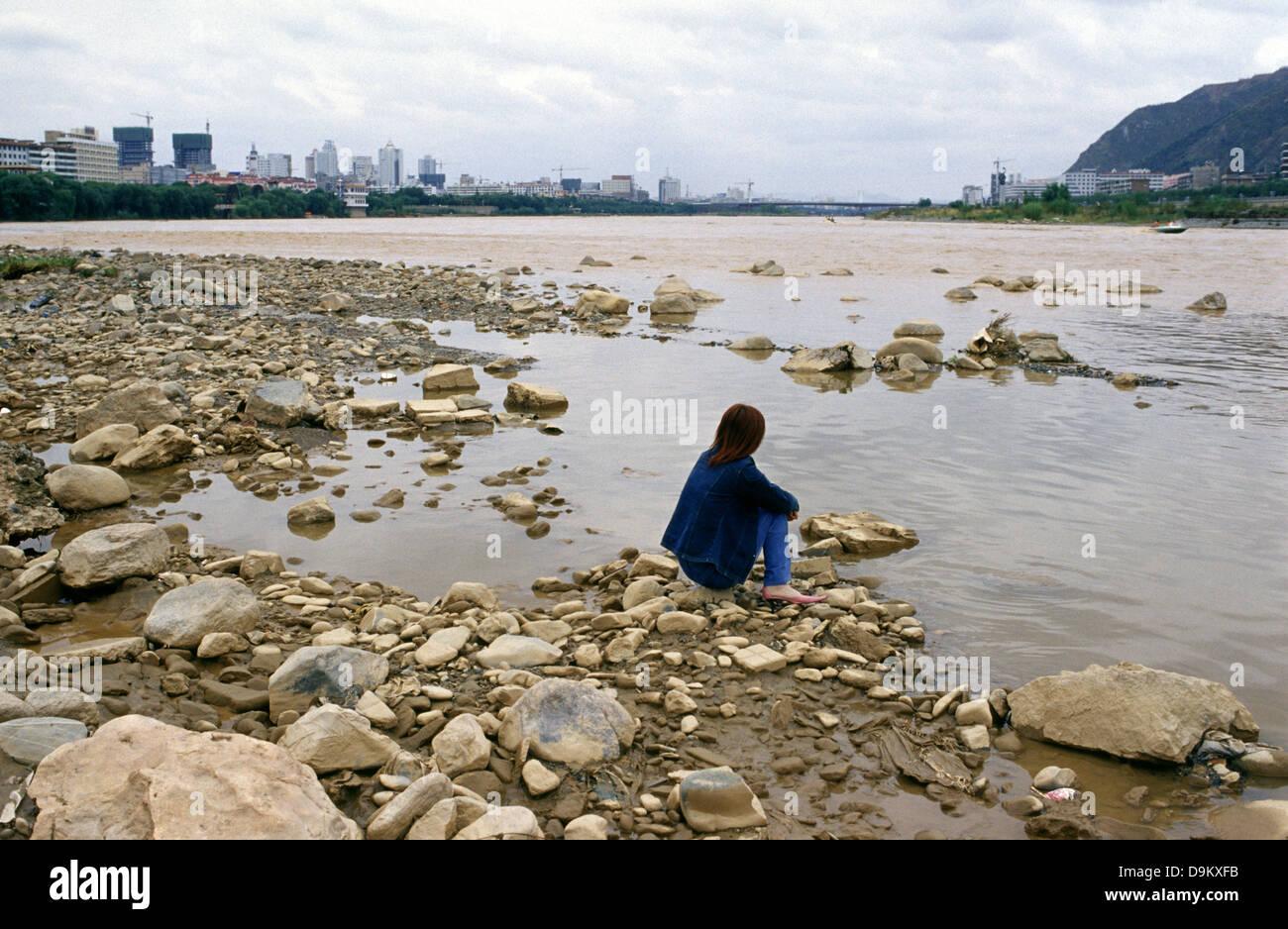 Lone young Chinese woman siting at the shore of the Huang He Yellow river flowing through Lanzhou city Gansu province China Stock Photo