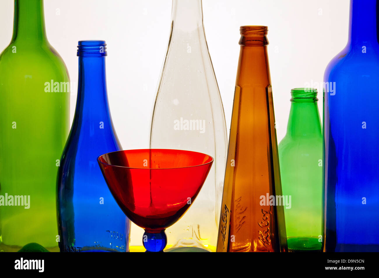 An abstract arrangement of multi-coloured glass bottles of different shapes and a red goblet against a backlit white screen Stock Photo