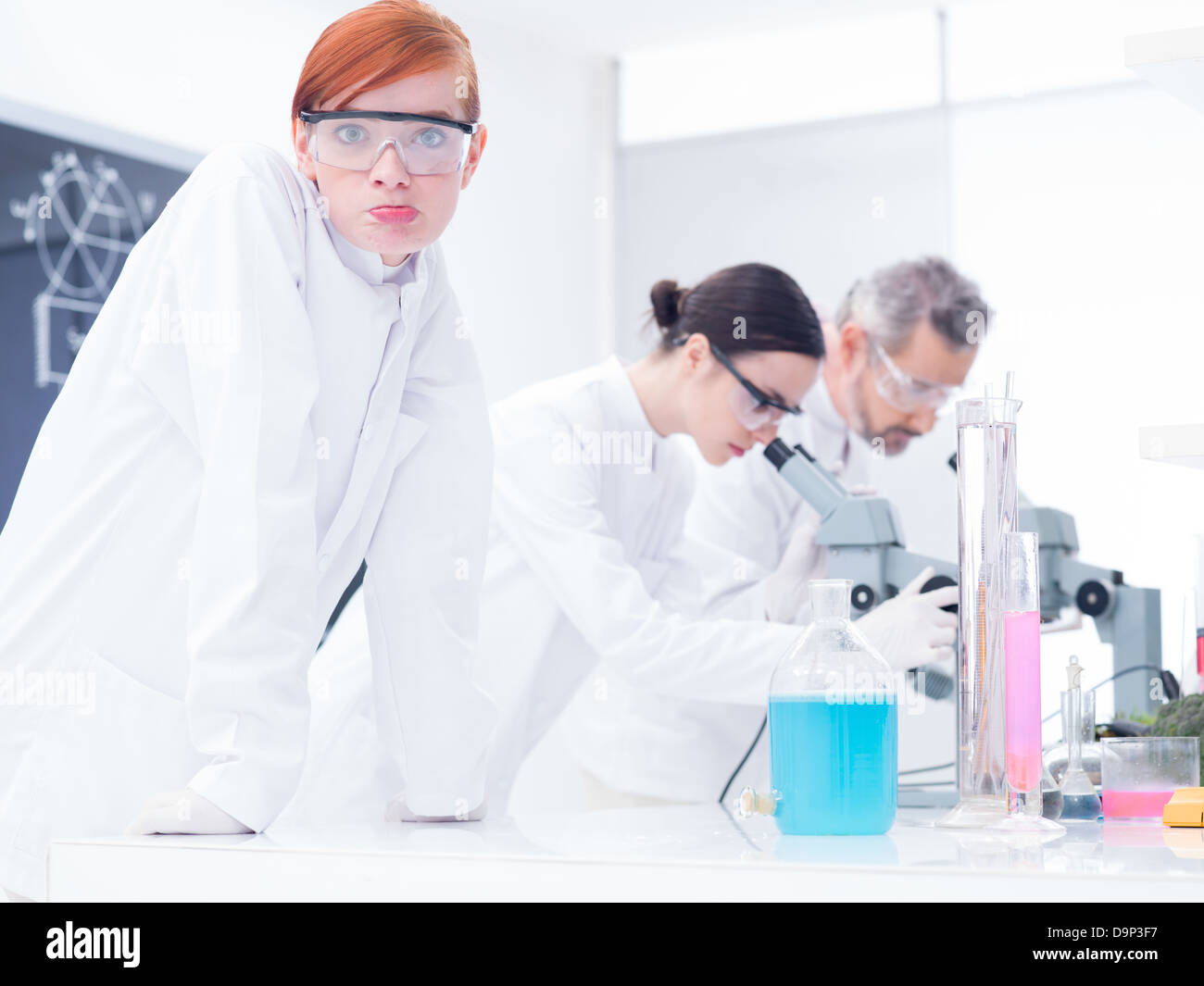 close-up of a beautiful girl student in a chemistry lab looking confused in the cameraand another two scientists analyzing under Stock Photo