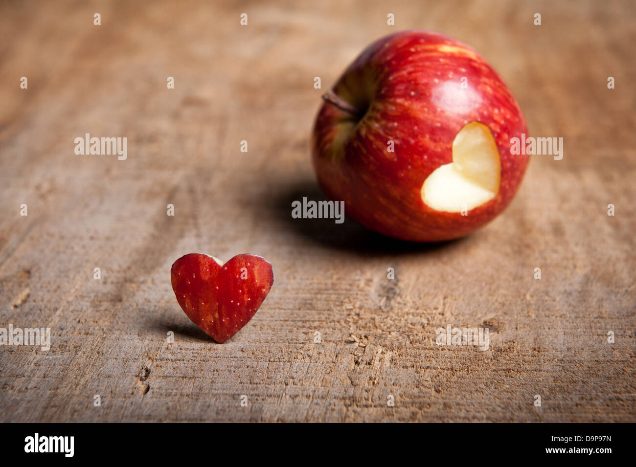 Red apple with heart-shape cut out. The cut-out heart is placed in front/next to the apple on a warm-toned wooden surface. Love Stock Photo