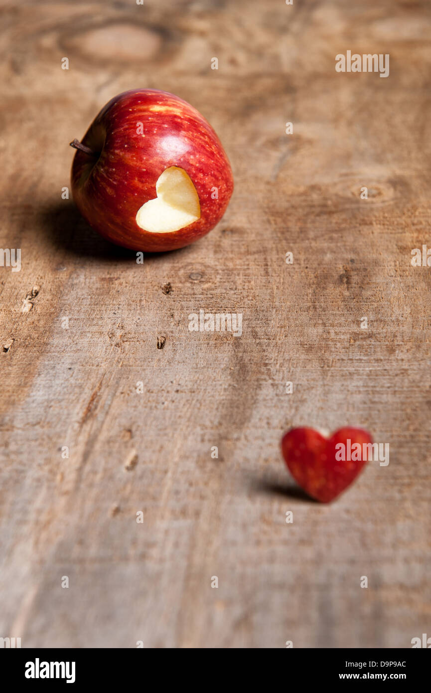 Red apple with heart-shape cut out. The cut-out heart is placed in front/next to the apple on a warm-toned wooden surface. Love Stock Photo