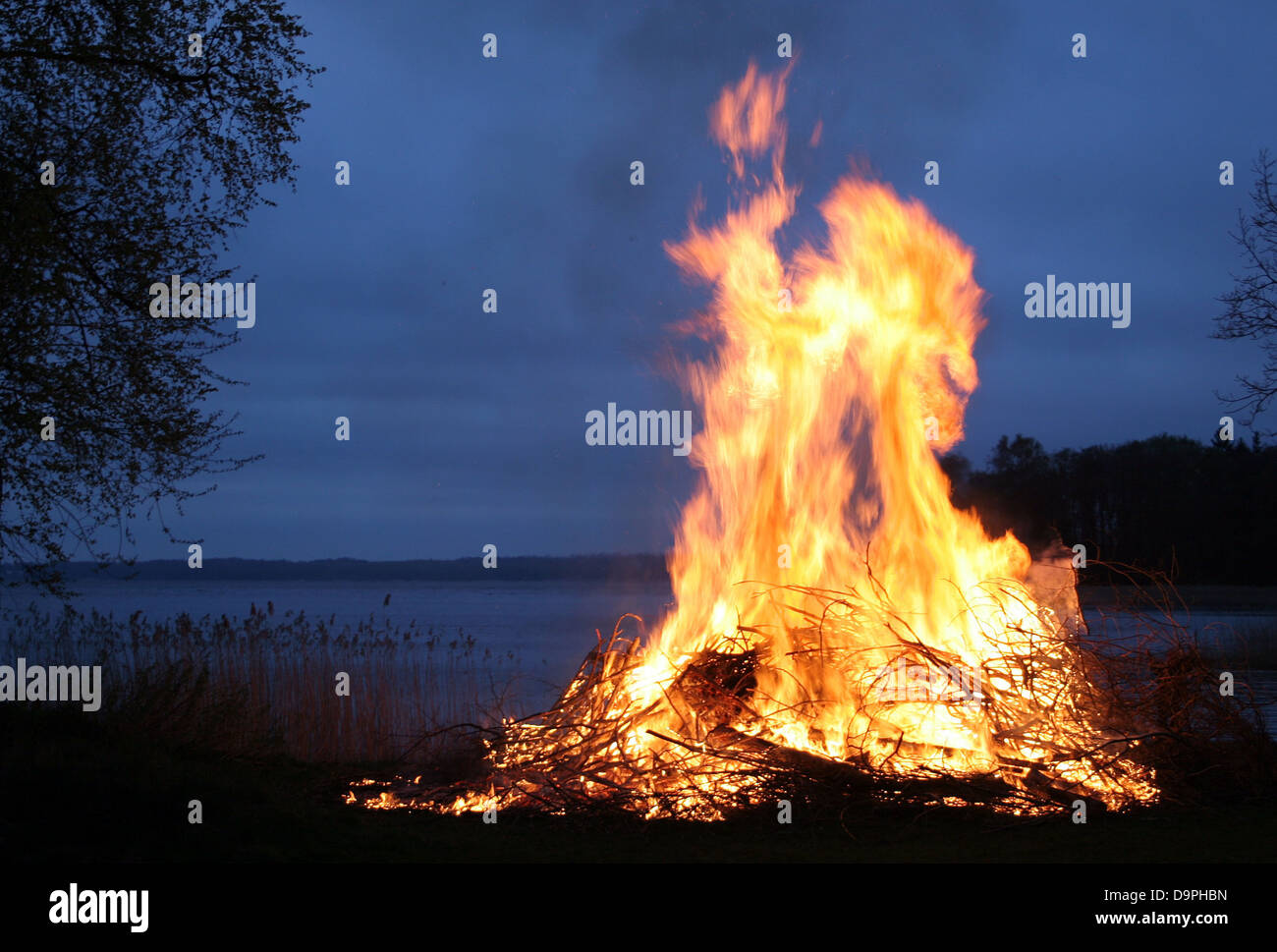 This scene depicts a bonfire in Sweden, surrounded by flames that light up the night sky. The flames create a striking contrast against the dark clouds, and the fire reflects in the nearby lake. The silhouettes of trees and nature add depth to the scene, emphasizing the beauty of the outdoors in the evening sky. Stock Photo