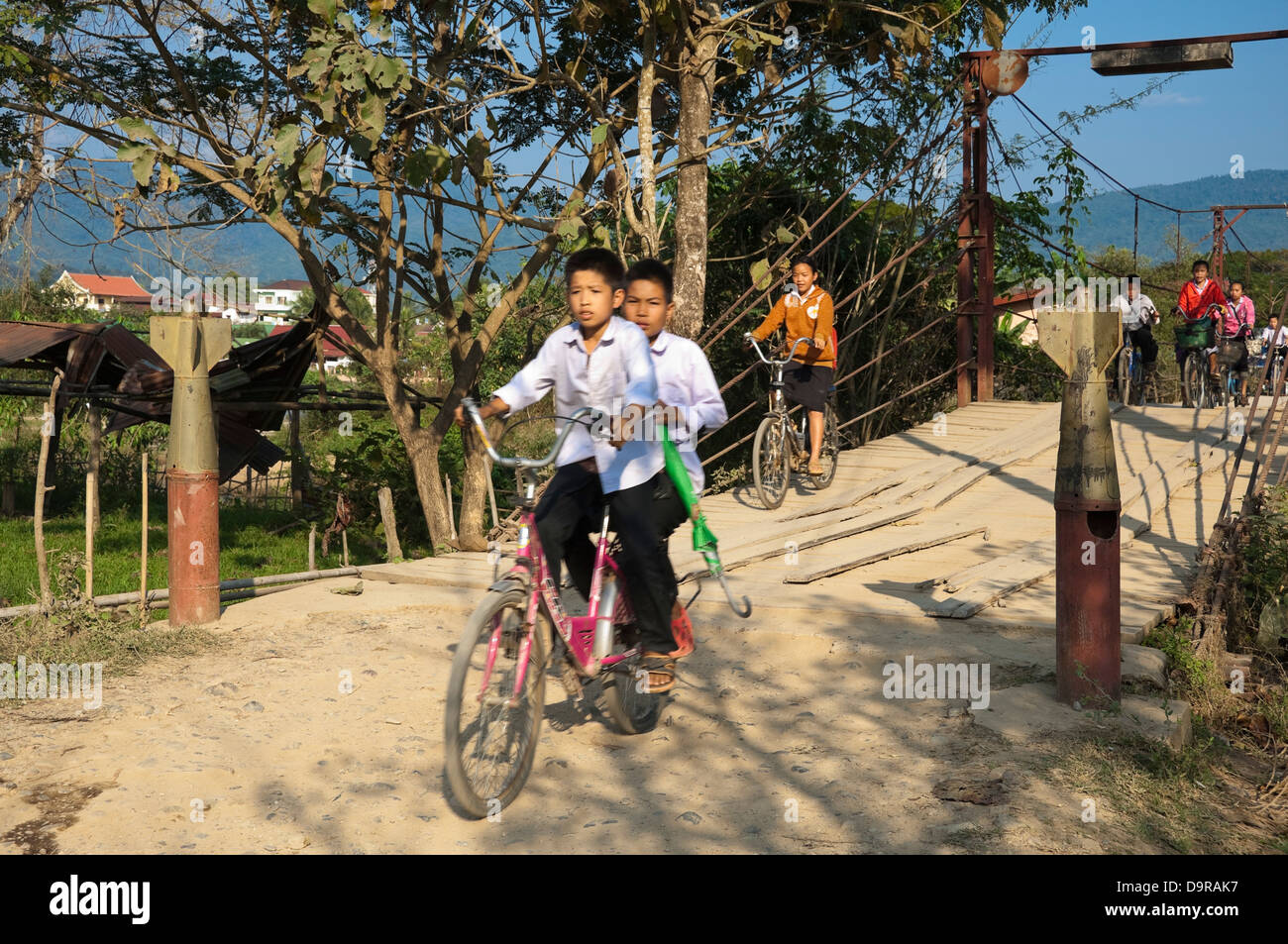 Horizontal portrait of young Laotian children riding on their bikes across a bridge with bomb casting bollards in Vang Vieng. Stock Photo