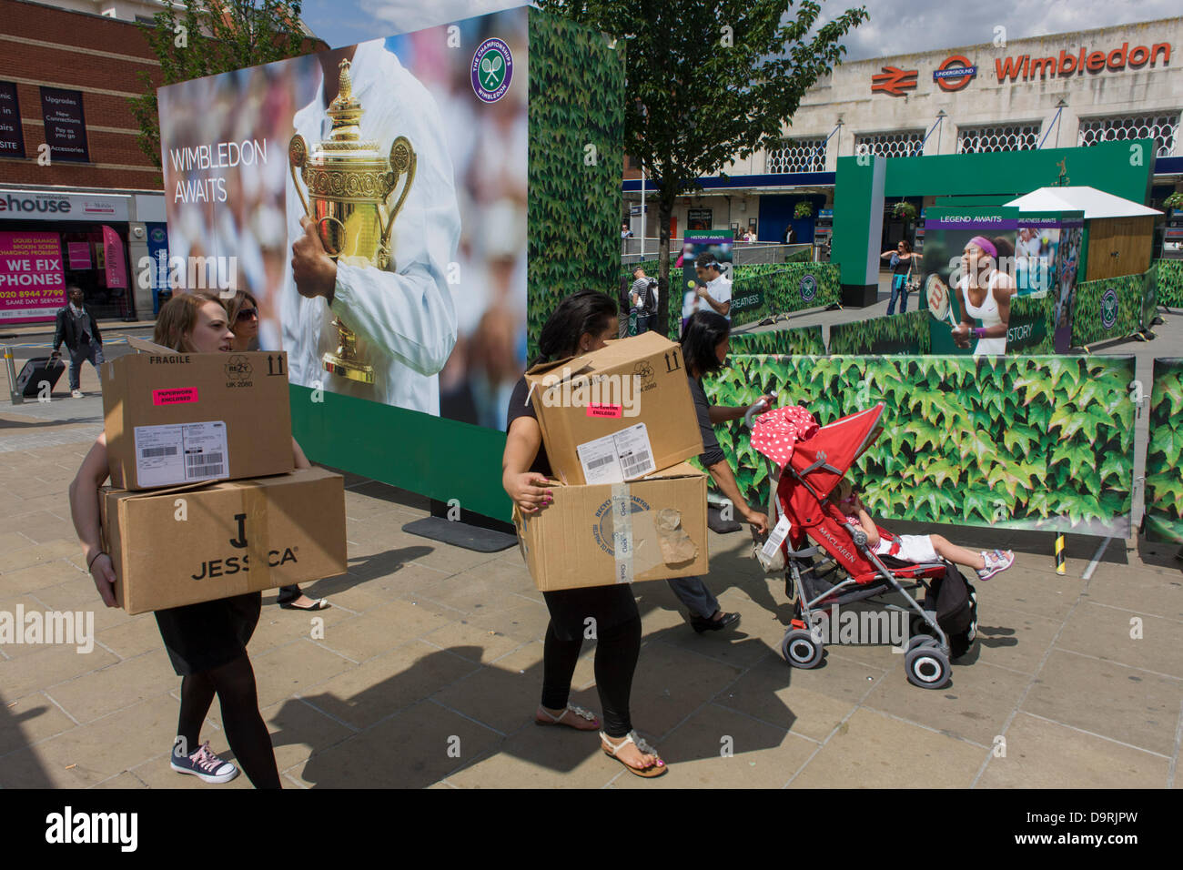 Wimbledon, England, 25th June 2013 - Day 2 of the annual lawn tennis championships women carry boxes and spectators mingle with locals near a large champions trophy billboard, outside the mainline and underground (subway) station in the south London suburb. The Wimbledon Championships, the oldest tennis tournament in the world, have been held at the nearby All England Club since 1877. Stock Photo