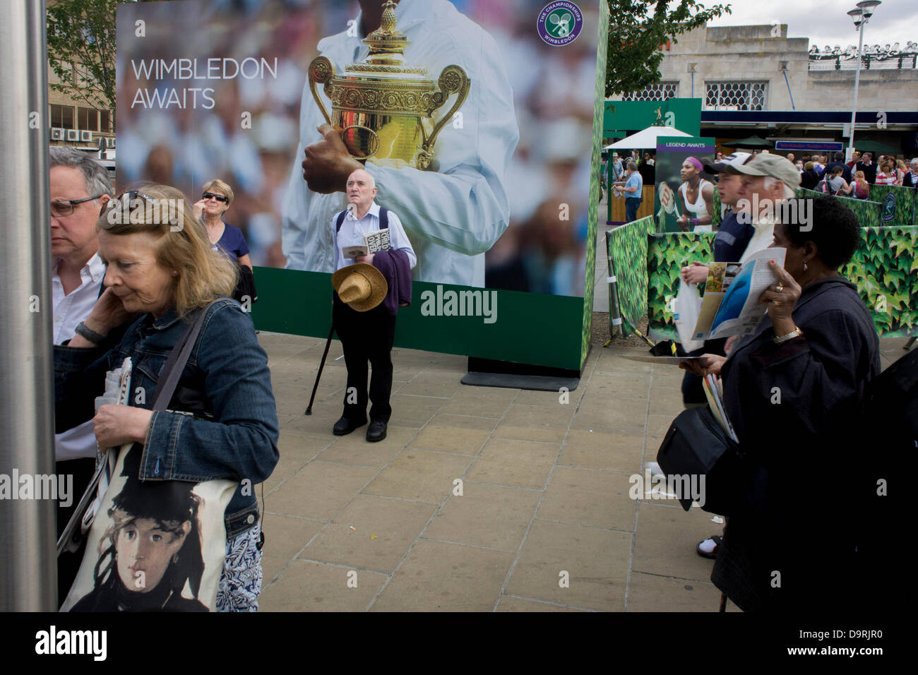 Wimbledon, England, 25th June 2013 - Day 2 of the annual lawn tennis championships and spectators mingle with locals near a large champions trophy billboard, outside the mainline and underground (subway) station in the south London suburb. The Wimbledon Championships, the oldest tennis tournament in the world, have been held at the nearby All England Club since 1877. Stock Photo