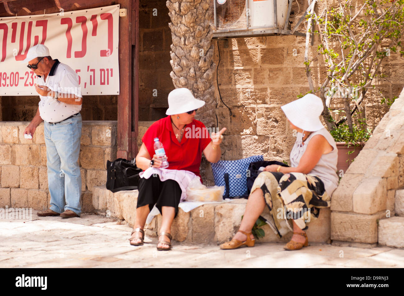 Israel Old Jaffa Yafo two 2 old elderly women ladies sitting shade chatting talking animated man tourists sun hat hats eating drinking Stock Photo